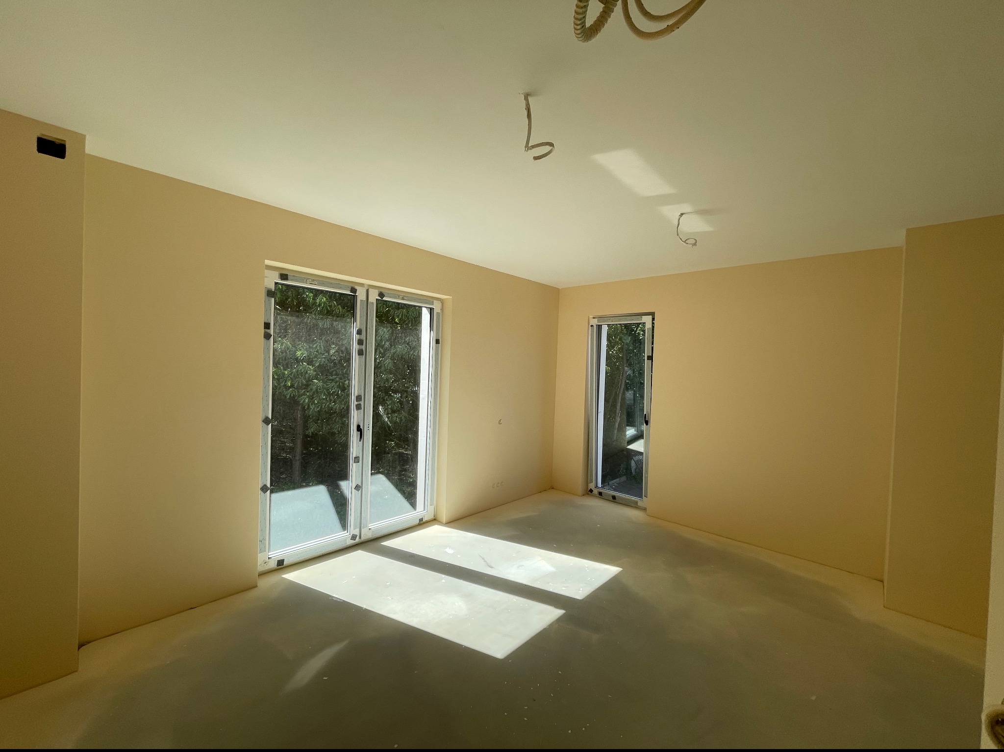 Pomalowane na beżowo wnętrze pokoju z dużymi oknami balkonowymi i jednym mniejszym oknem, widoczne kable wystające z sufitu, podłoga zabezpieczona folią malarską.