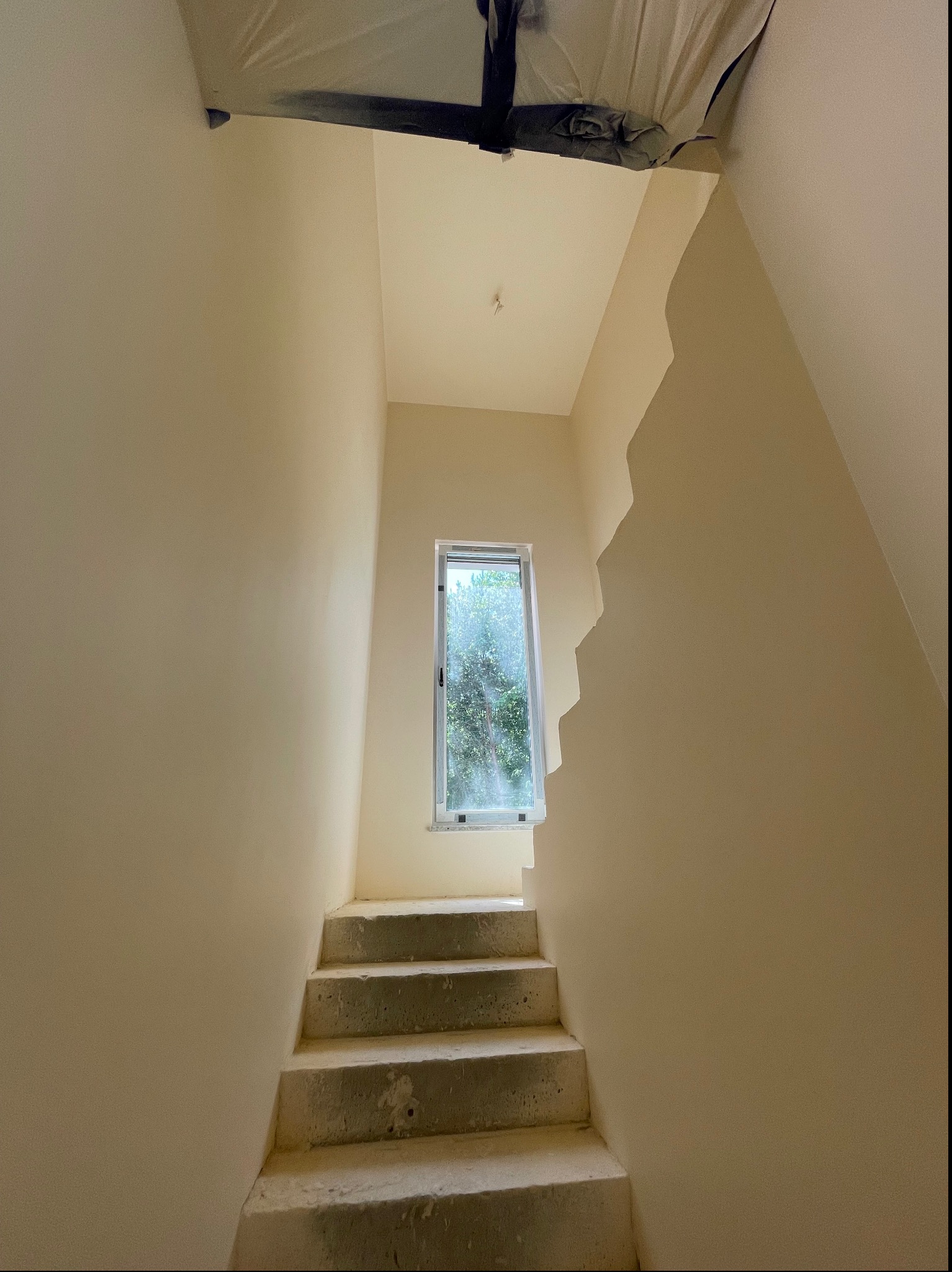 Klatka schodowa z betonowymi schodami po malowaniu ścian na jasny kremowy kolor, okno z widokiem na zieleń w tle, zabezpieczenie folią malarską widoczne pod sufitem.