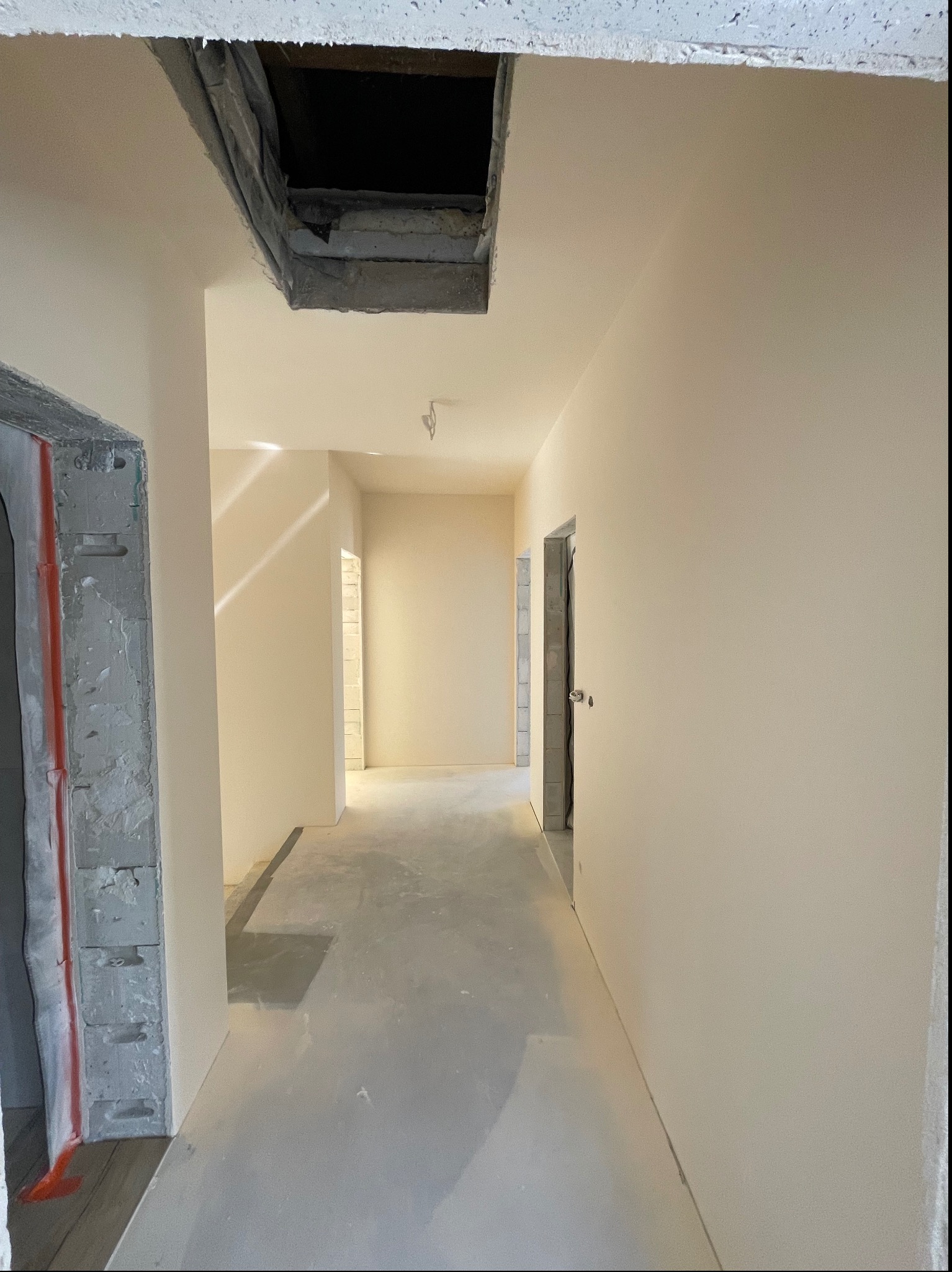 Korytarz w stanie surowym po szpachlowaniu i gruntowaniu ścian na jasny odcień, widoczne niedokończone ościeżnice i otwór w suficie.