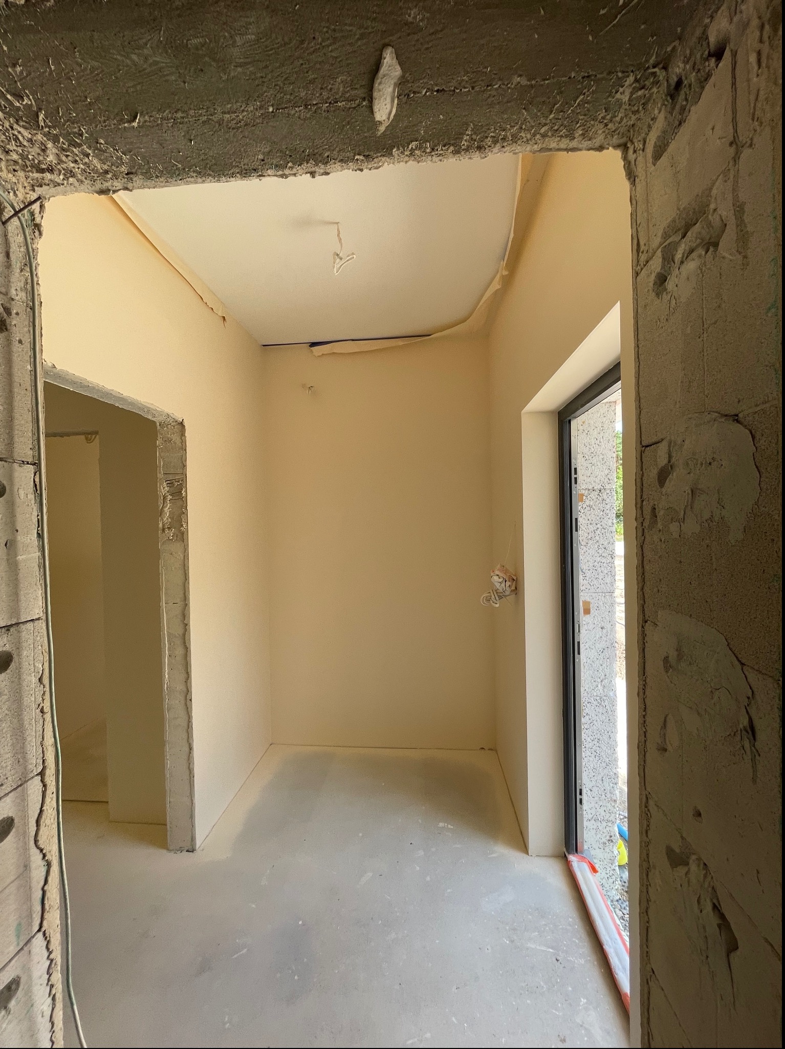 Wnętrze w trakcie remontu: świeżo wyszpachlowane i pomalowane ściany na beżowo, surowy beton wokół futryn drzwiowych i ościeżnic, przygotowane do dalszych prac wykończeniowych.