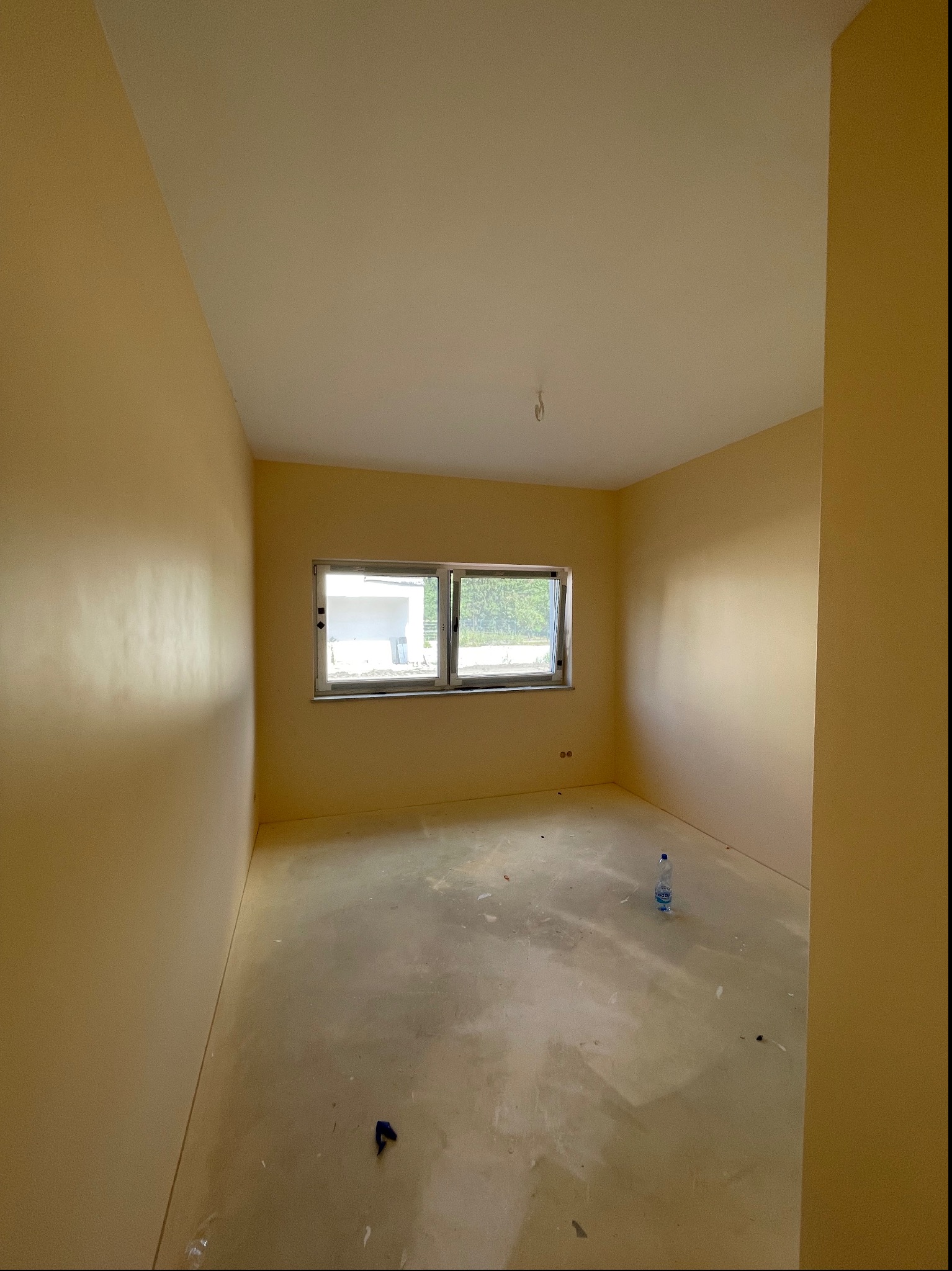 Pomalowane na jasnożółto puste pomieszczenie z oknem, na podłodze widoczna butelka wody i drobne zabrudzenia po remoncie.