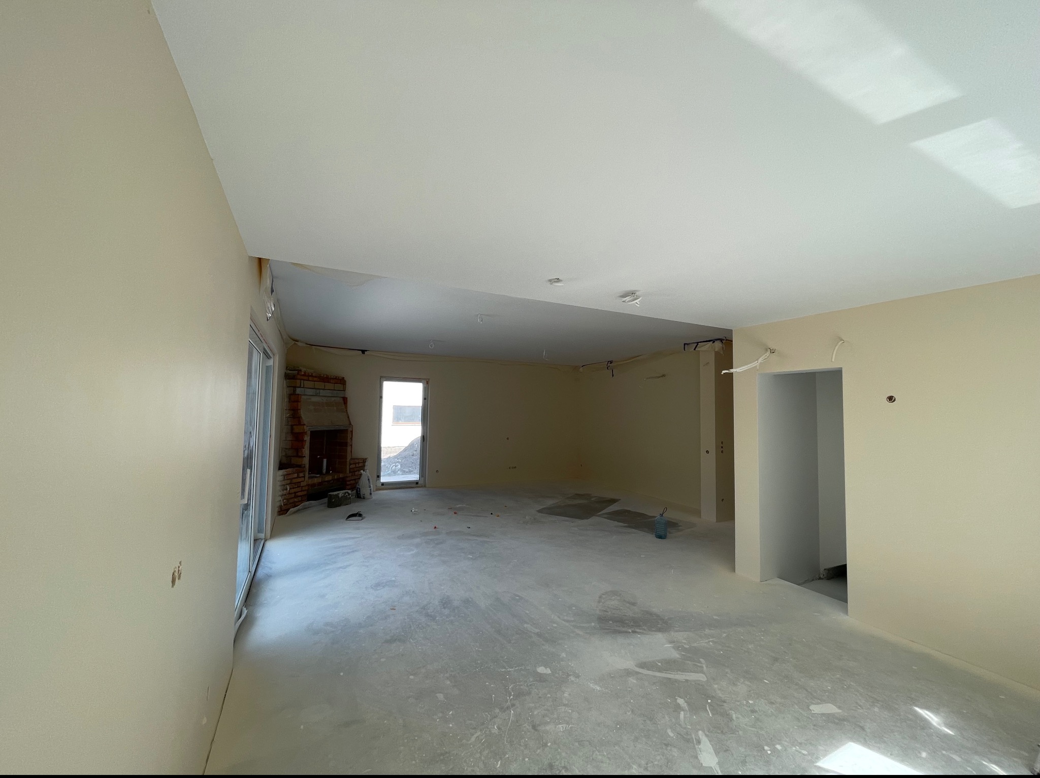 Wnętrze w trakcie remontu: świeżo pomalowane ściany w odcieniach beżu, surowy beton na podłodze z widocznymi śladami prac, ceglany kominek w tle, przygotowane do dalszych prac wykończeniowych.