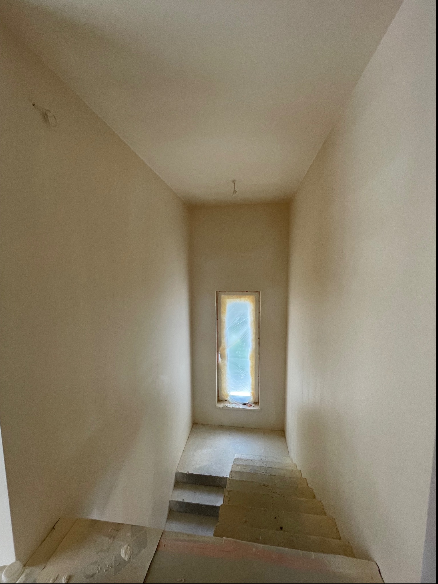Wnętrze klatki schodowej w trakcie prac wykończeniowych: ściany pokryte świeżo nałożoną gładzią szpachlową, okno zabezpieczone folią malarską, widoczne schody z resztkami materiałów budowlanych.