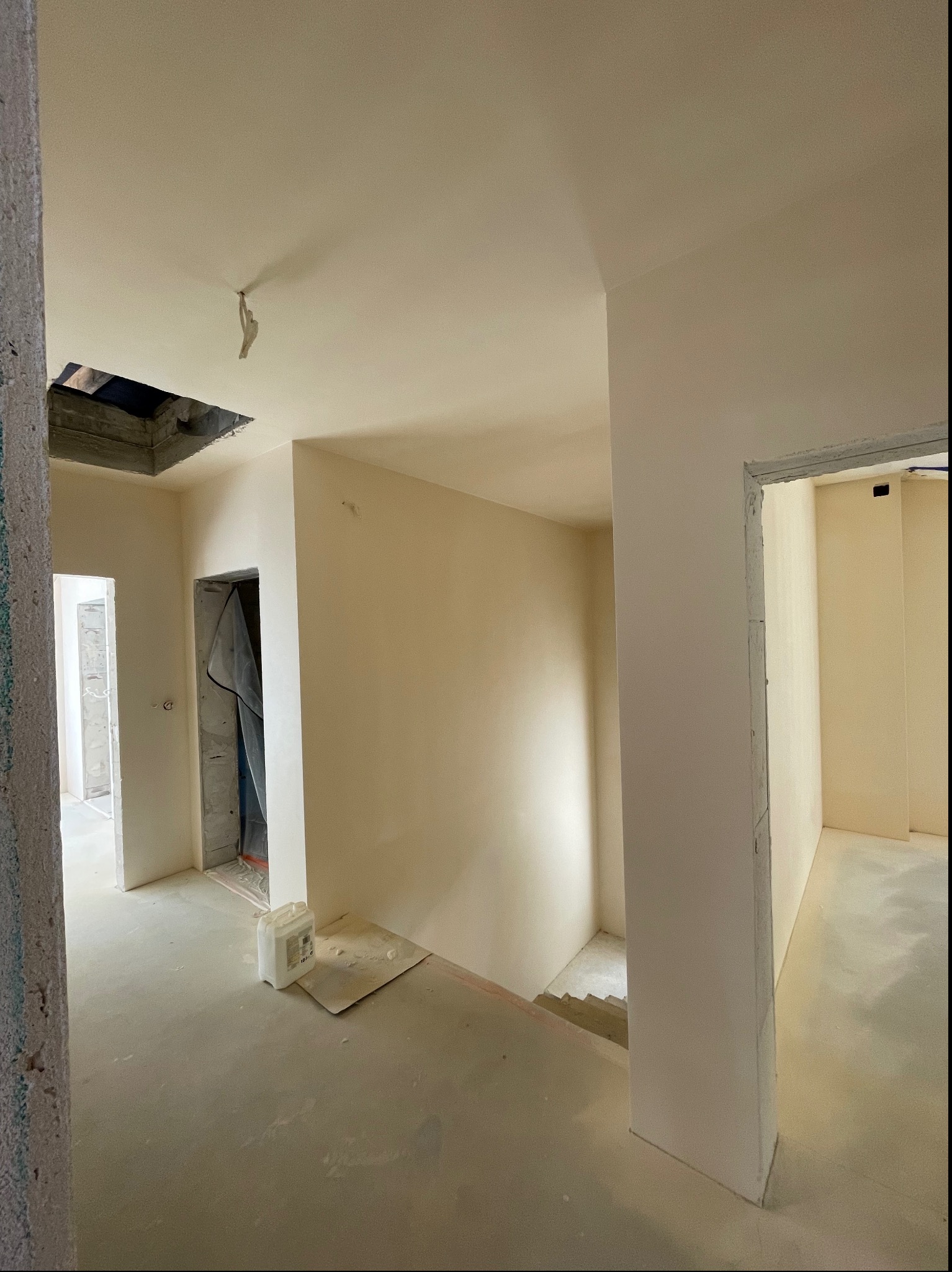 Wnętrze w trakcie prac wykończeniowych: świeżo pomalowane ściany w odcieniu beżu, surowy beton na podłodze, widoczne otwory w suficie, zabezpieczone folią ościeżnice drzwiowe, oraz kanister...