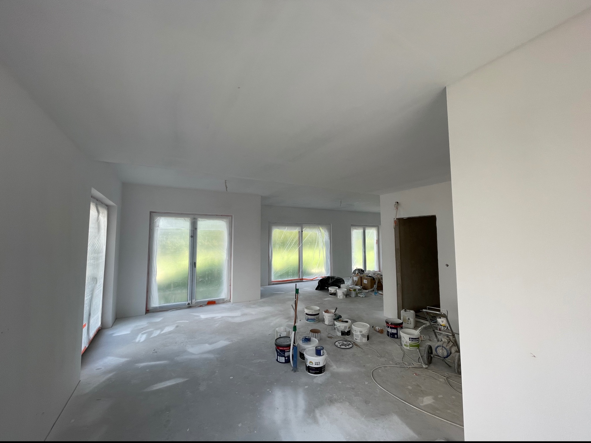 Wnętrze w trakcie prac wykończeniowych: świeżo pomalowane na biało ściany i sufit, okna zabezpieczone folią, widoczne puszki z farbą i sprzęt malarski na podłodze pokrytej plamami farby.