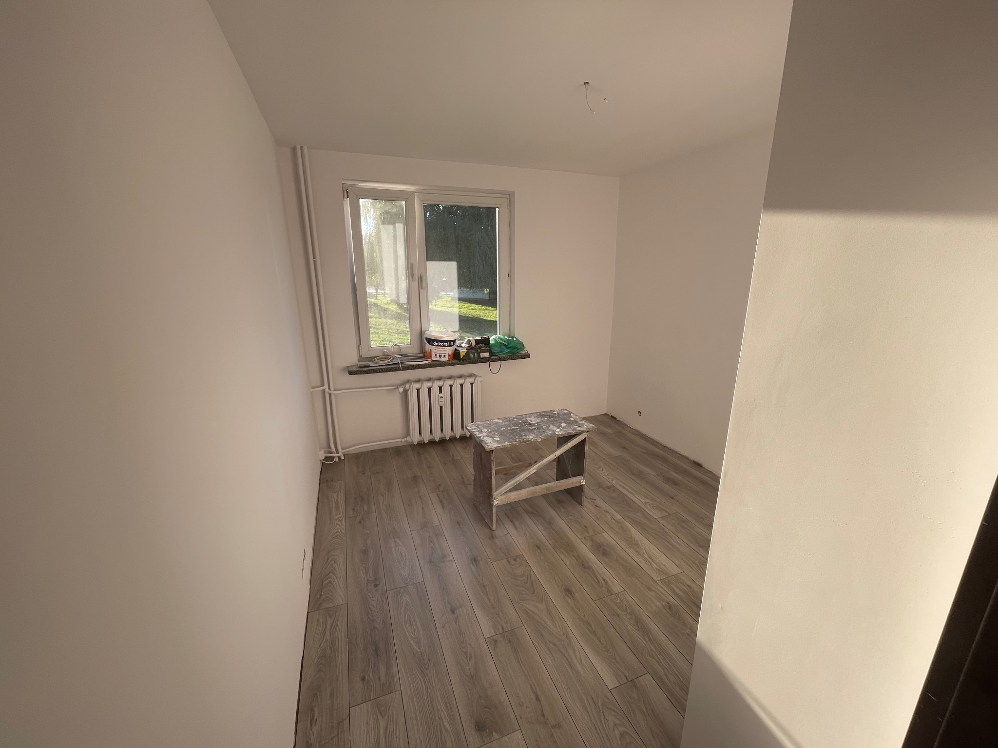 Wnętrze pokoju po malowaniu: białe ściany, drewniana podłoga, okno z widokiem na zewnątrz, drabinka malarska. Na parapecie puszki z farbą. Minimalistyczny efekt.