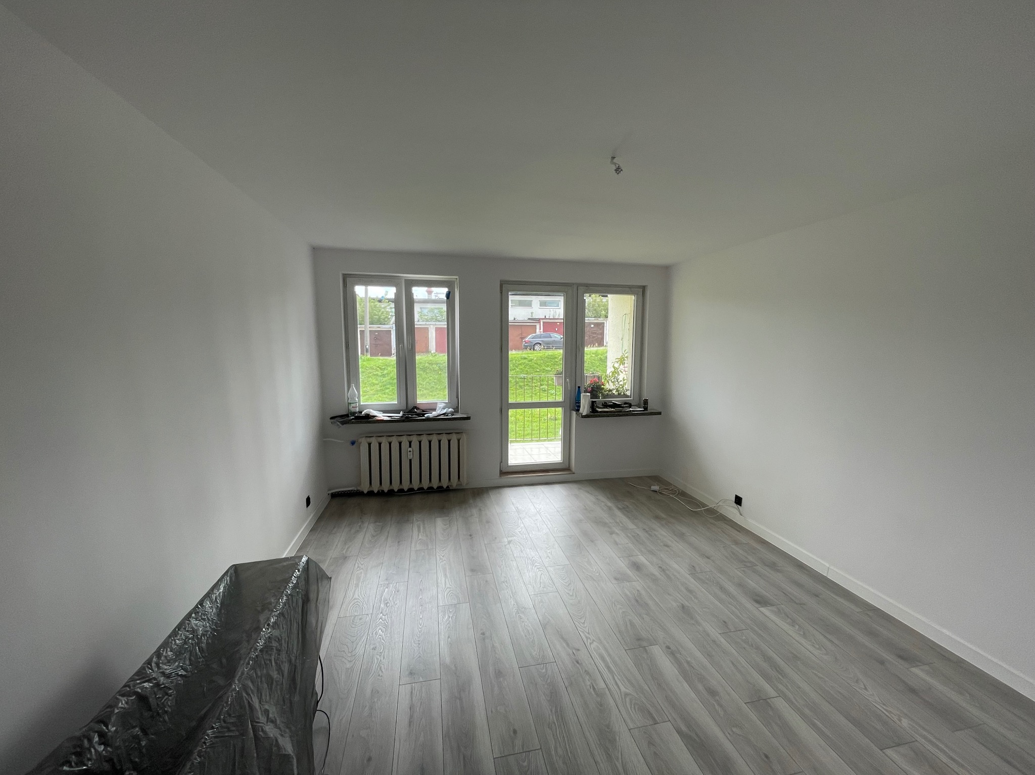 Jasne, odświeżone wnętrze po malowaniu: białe ściany, szara podłoga. Okno z widokiem na zieleń. Grzejnik pod oknem. Meble zabezpieczone folią malarską.