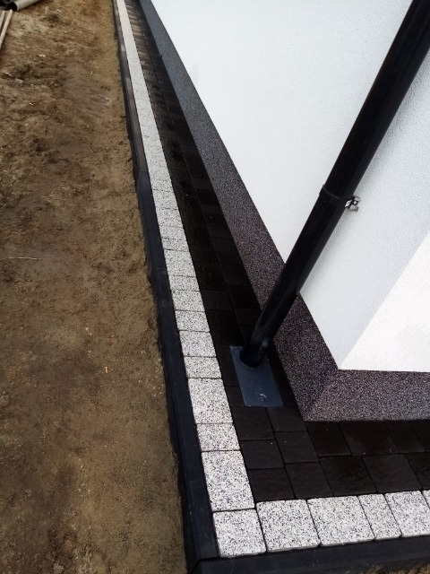 Ułożona kostka brukowa w Aleksandrowie, obramowanie budynku z rurą spustową, widoczne różne odcienie szarości i czerni kostki.