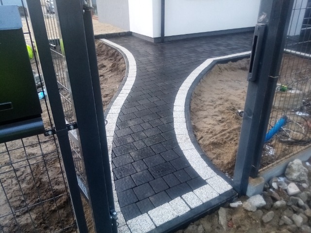 Ułożona ścieżka z ciemnej kostki brukowej obramowana jasną kostką, widok przez metalowe ogrodzenie w kierunku białego budynku.