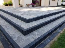 Schody wejściowe wyłożone szarą kostką brukową z ciemnymi obrzeżami, prowadzące do jasnego domu z kolumnami i kwiatami w donicach.