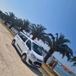 Biały van Ford Transit ustawiony ukośnie na piaszczystej drodze, w tle palmy, błękitne niebo i woda z zacumowanymi łodziami.