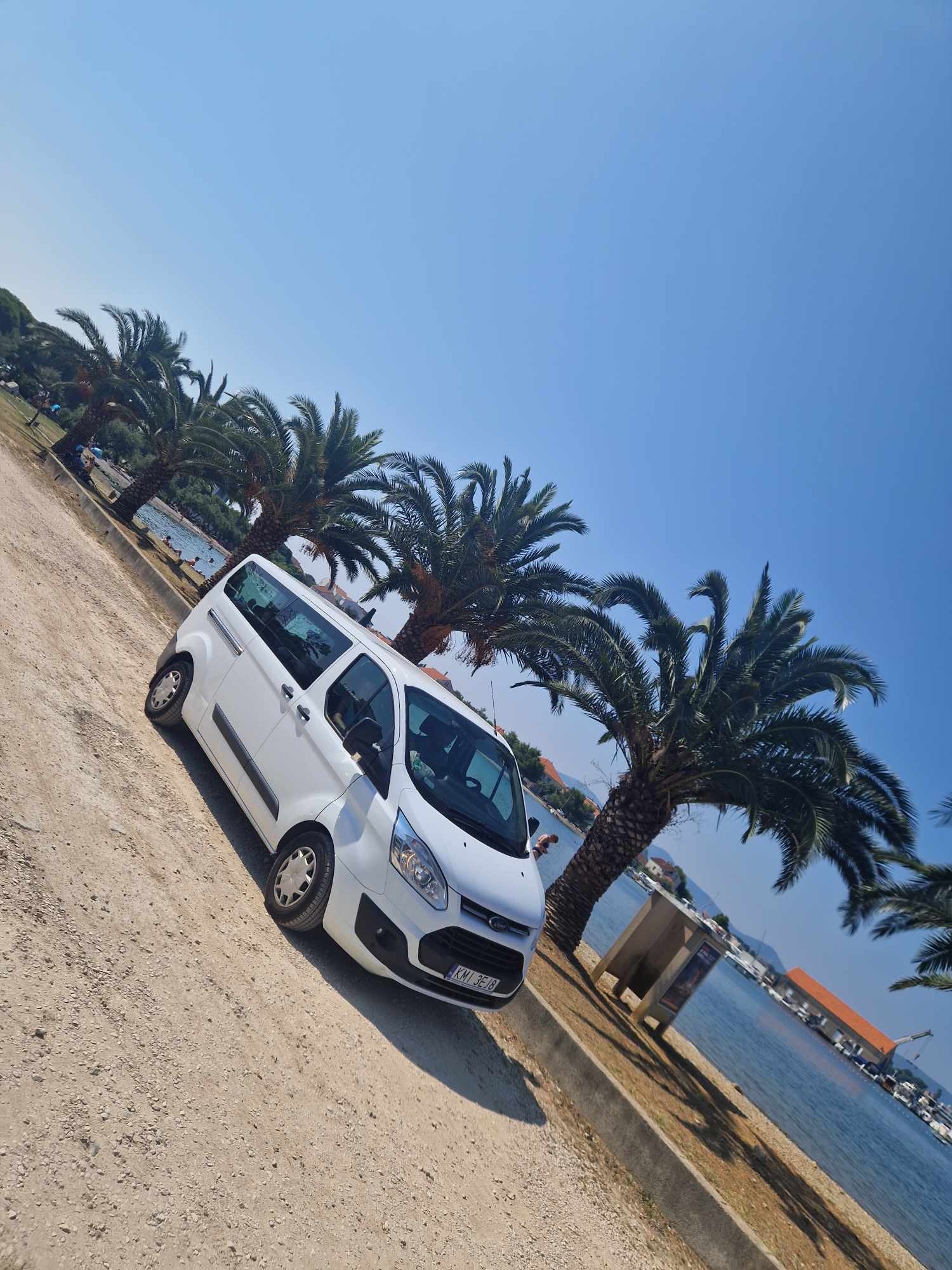 Biały van Ford Transit ustawiony ukośnie na piaszczystej drodze, w tle palmy, błękitne niebo i woda z zacumowanymi łodziami.