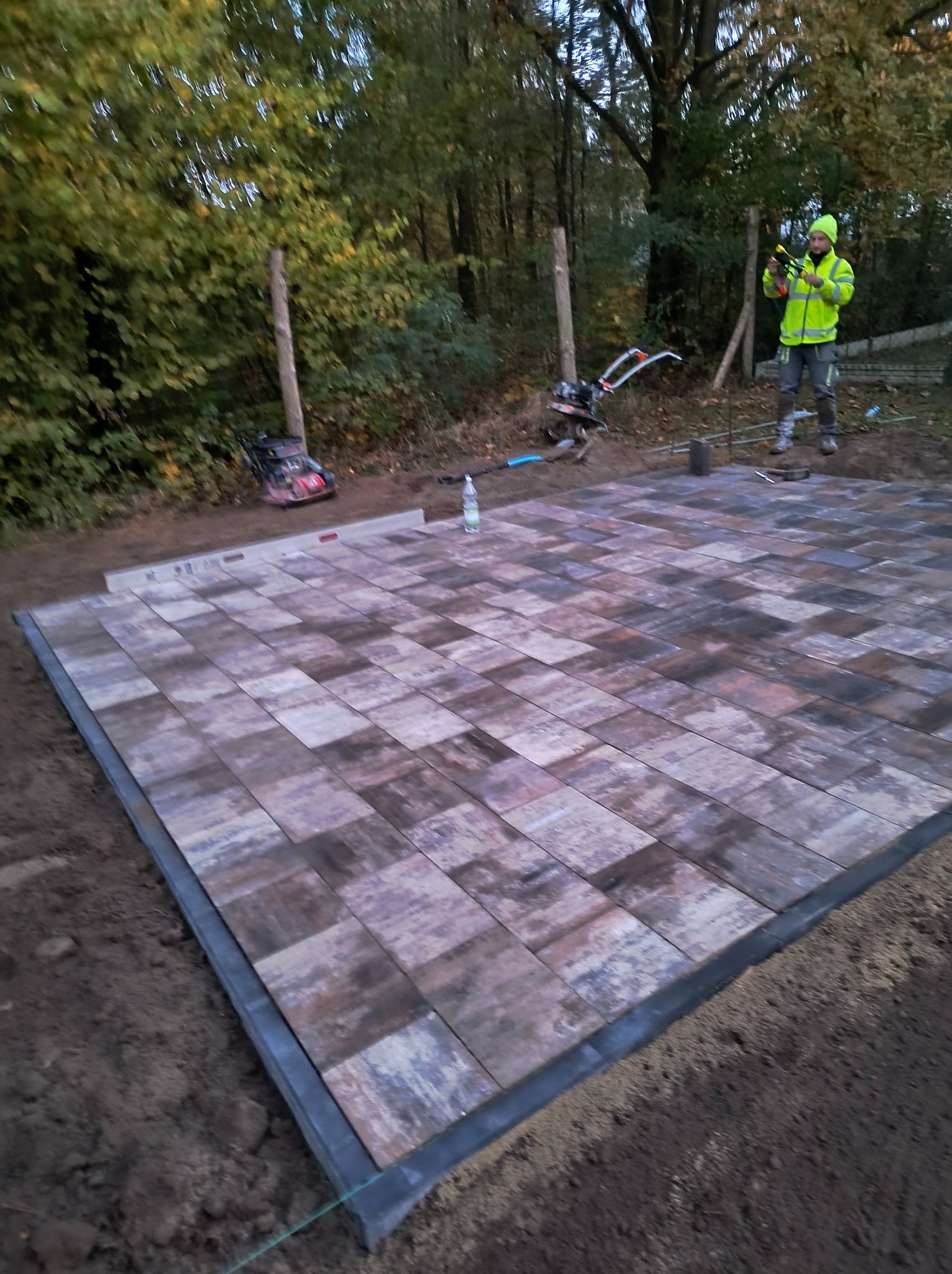 Ułożona część powierzchni z kostki brukowej w odcieniach szarości i brązu, widoczny pracownik w odblaskowej kurtce, poziomica, zagęszczarka i butelka wody mineralnej.