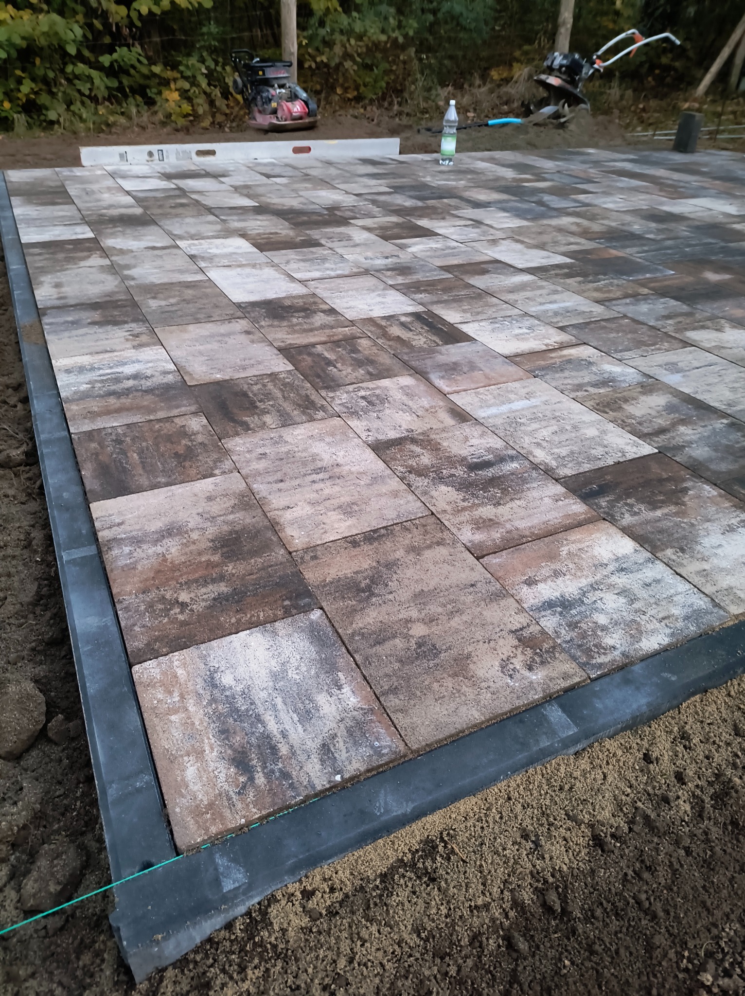 Ułożona nawierzchnia z kostki brukowej o nieregularnym wzorze, obramowana czarnym obrzeżem, widoczny niwelator laserowy, zagęszczarka i butelka wody w tle.