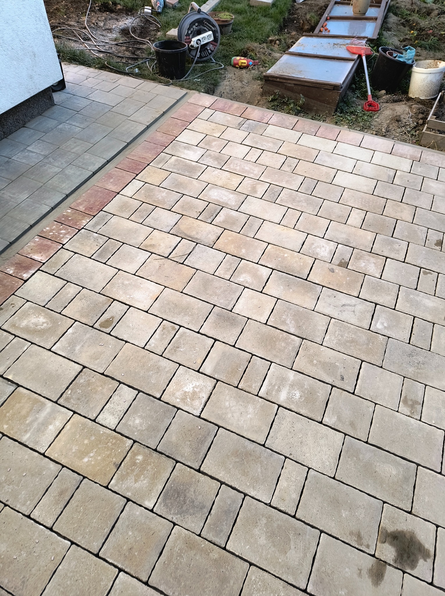 Ułożona kostka brukowa w odcieniach beżu i szarości, obrzeże z czerwonej kostki, widoczne narzędzia ogrodowe i akcesoria w tle.