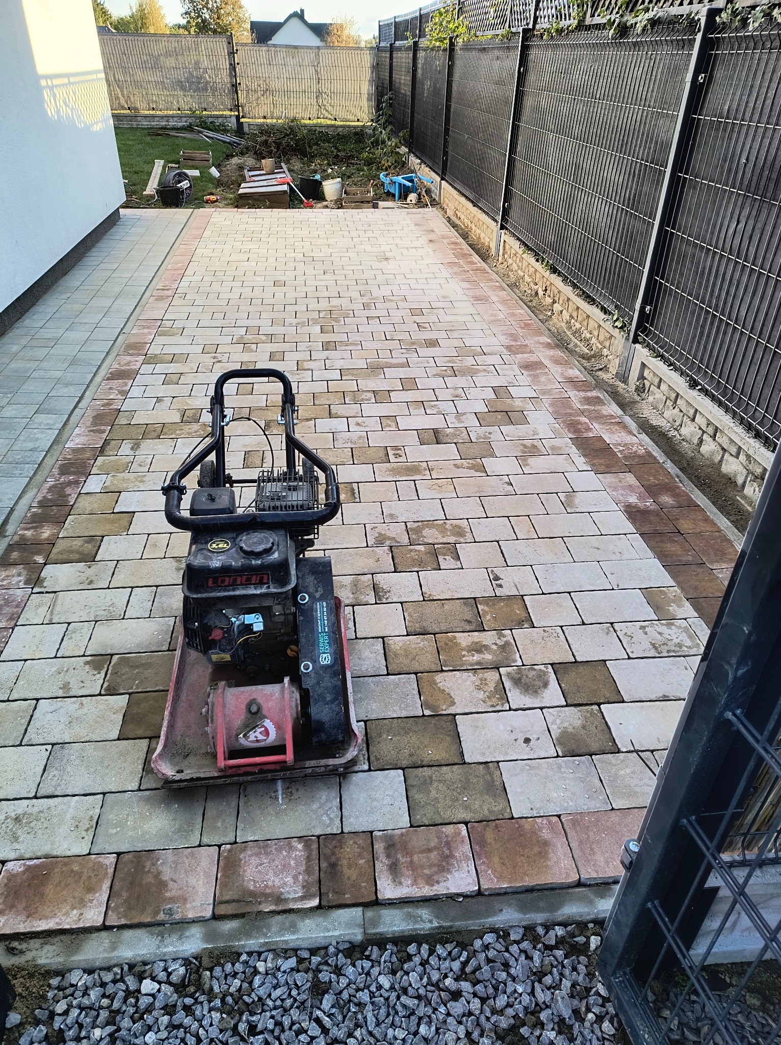 Świeżo ułożona aleja z kostki brukowej w odcieniach beżu i brązu, częściowo utwardzona wibratorem spalinowym, widoczny czarny metalowy płot i przygotowane podłoże pod dalszą część alei.