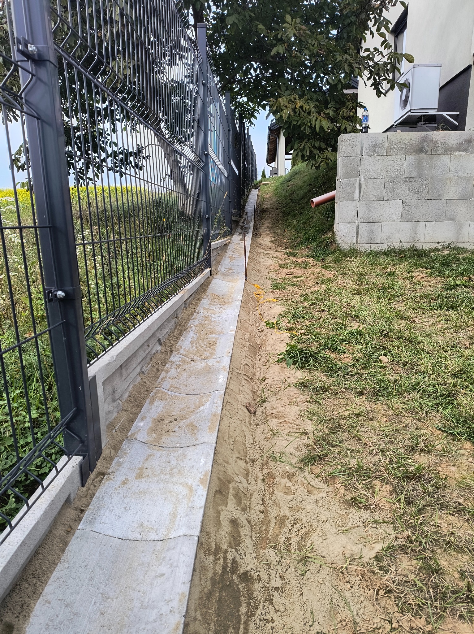 Betonowy rów odwadniający, częściowo zasypany piaskiem, biegnący wzdłuż ogrodzenia panelowego w Lublinie, z widocznym budynkiem mieszkalnym w tle.