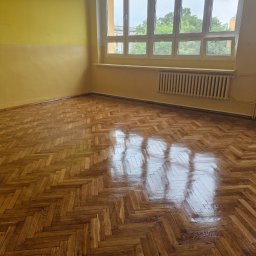 Felidwood usługi parkieciarskie - Wnętrze pokoju z parkietem ułożonym w jodełkę, odbijającym światło z okna. Ściany pomalowane na żółto, widoczne grzejniki pod oknem.