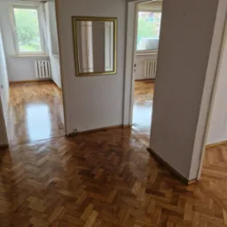 Błyszcząca, odnowiona podłoga parkietowa ułożona w jodełkę, widoczna z perspektywy wejścia do pomieszczenia, z ramką lustra na ścianie w tle.