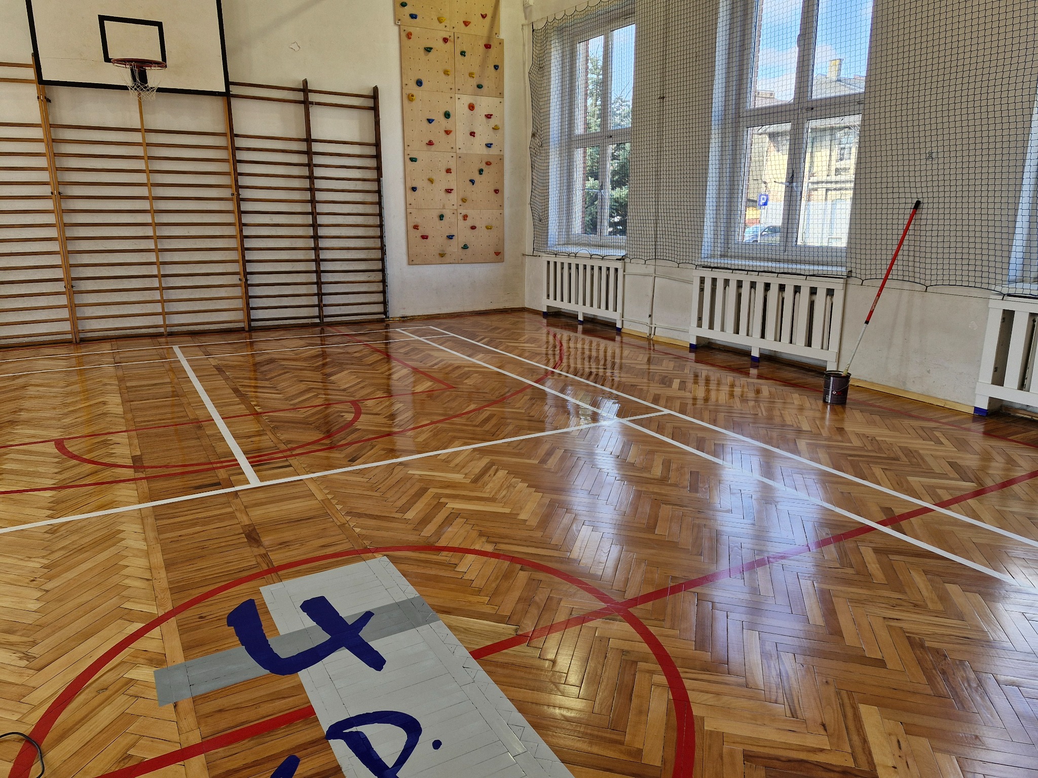 Sala gimnastyczna z parkietem ułożonym w jodełkę, widoczne linie boiska, drabinki gimnastyczne, ścianka wspinaczkowa i okna z siatką ochronną.