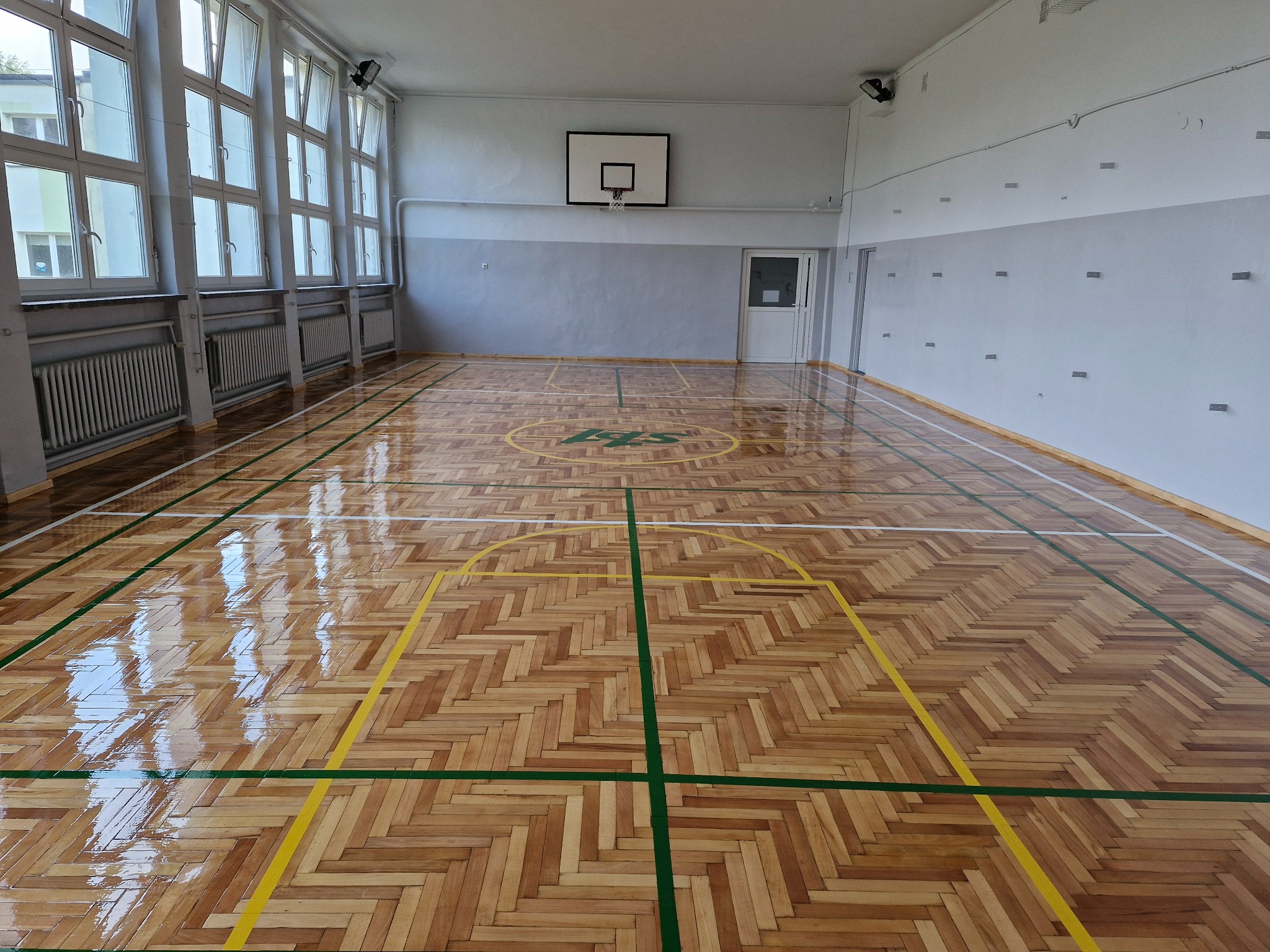 Błyszcząca, odnowiona podłoga z parkietu w sali gimnastycznej z widocznymi liniami boiska w kolorach żółtym i zielonym oraz herbem szkoły na środku.