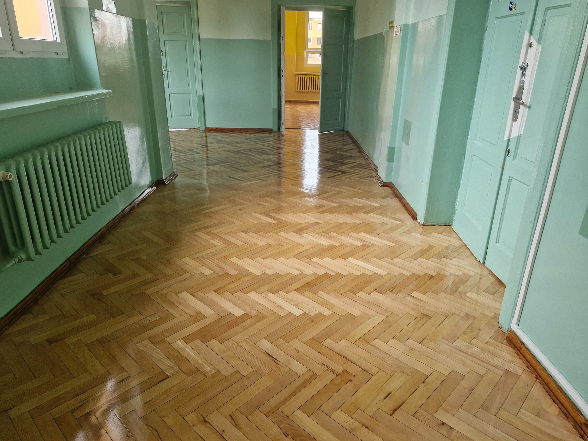 Świeżo wycyklinowany parkiet w jodełkę w korytarzu w Zawierciu, połyskujący lakier, widoczne drzwi w kolorze miętowym i grzejnik.