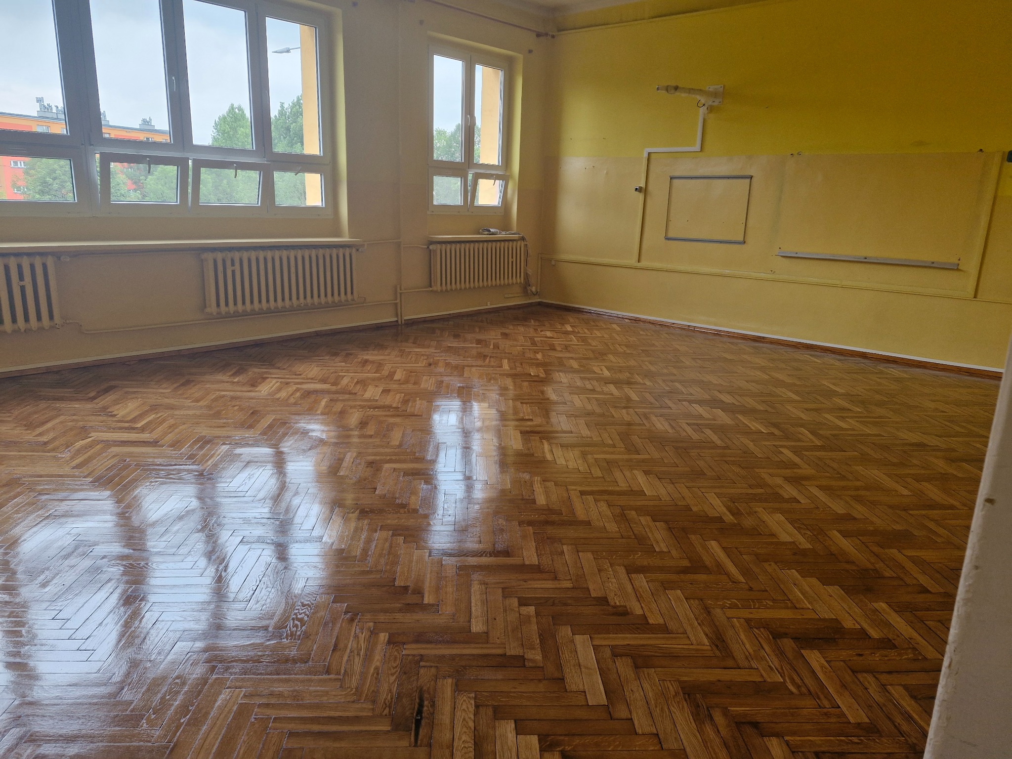 Wypolerowany parkiet ułożony w jodełkę w pustym, słonecznym pokoju z żółtymi ścianami i grzejnikami pod oknami.