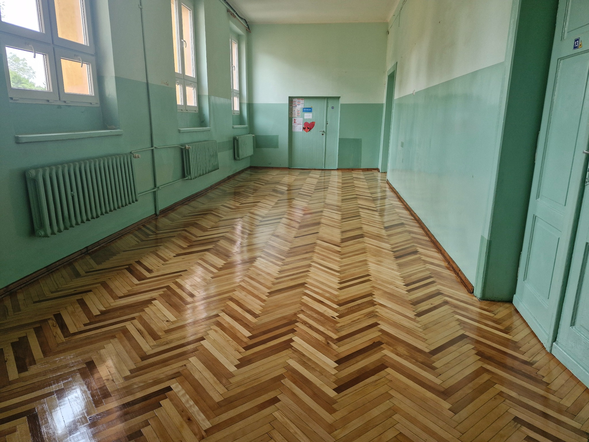 Wnętrze korytarza z parkietem ułożonym w jodełkę, ściany pomalowane na zielono z białym pasem u góry, widoczne grzejniki i drzwi.