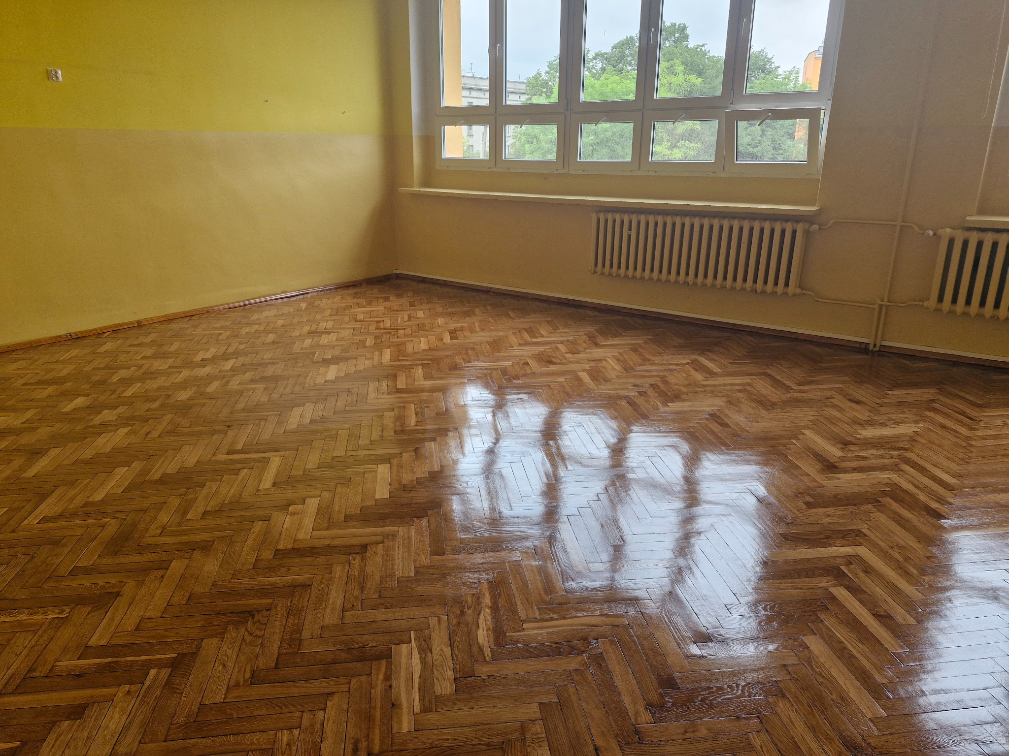 Wnętrze pokoju z parkietem ułożonym w jodełkę, odbijającym światło z okna. Ściany pomalowane na żółto, widoczne grzejniki pod oknem.
