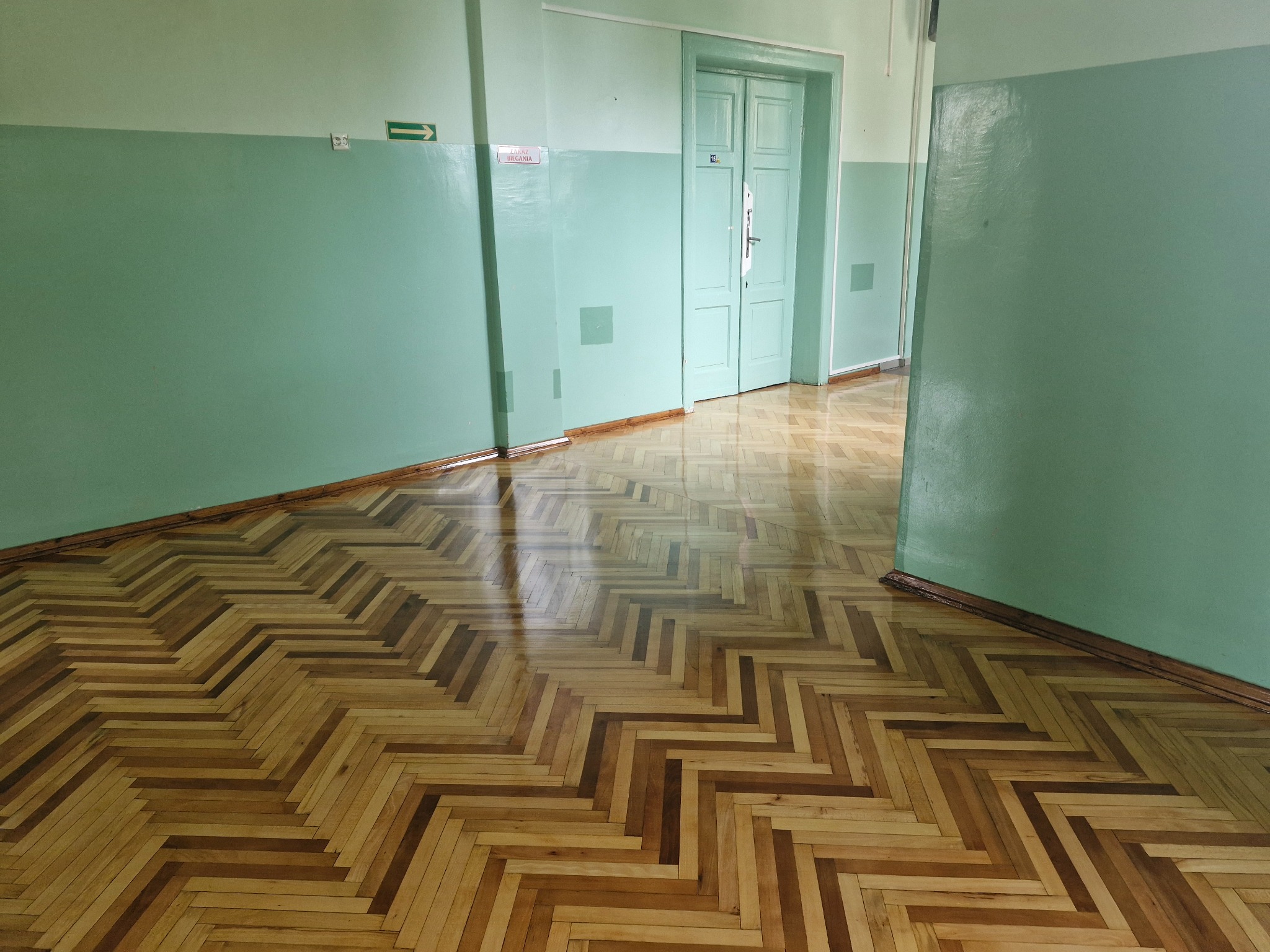 Błyszcząca, odnowiona podłoga parkietowa ułożona we wzór jodełki w korytarzu z zielonymi ścianami i otwartymi drzwiami w tle.