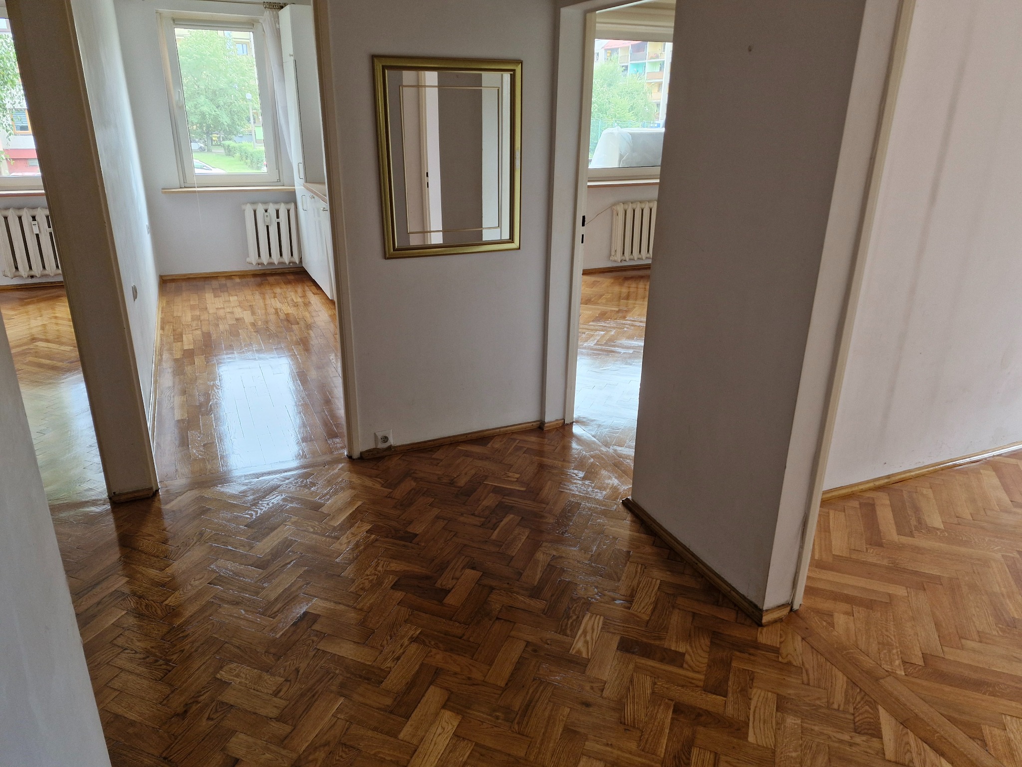 Błyszcząca, odnowiona podłoga parkietowa ułożona w jodełkę, widoczna z perspektywy wejścia do pomieszczenia, z ramką lustra na ścianie w tle.