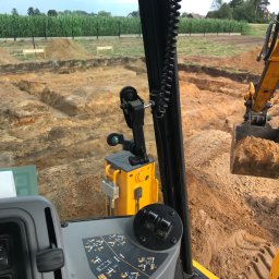 Kar-Geo-Bud - Widok z kabiny koparki na wykop pod fundamenty. Żółta maszyna w trakcie pracy na placu budowy. W tle pole kukurydzy.
