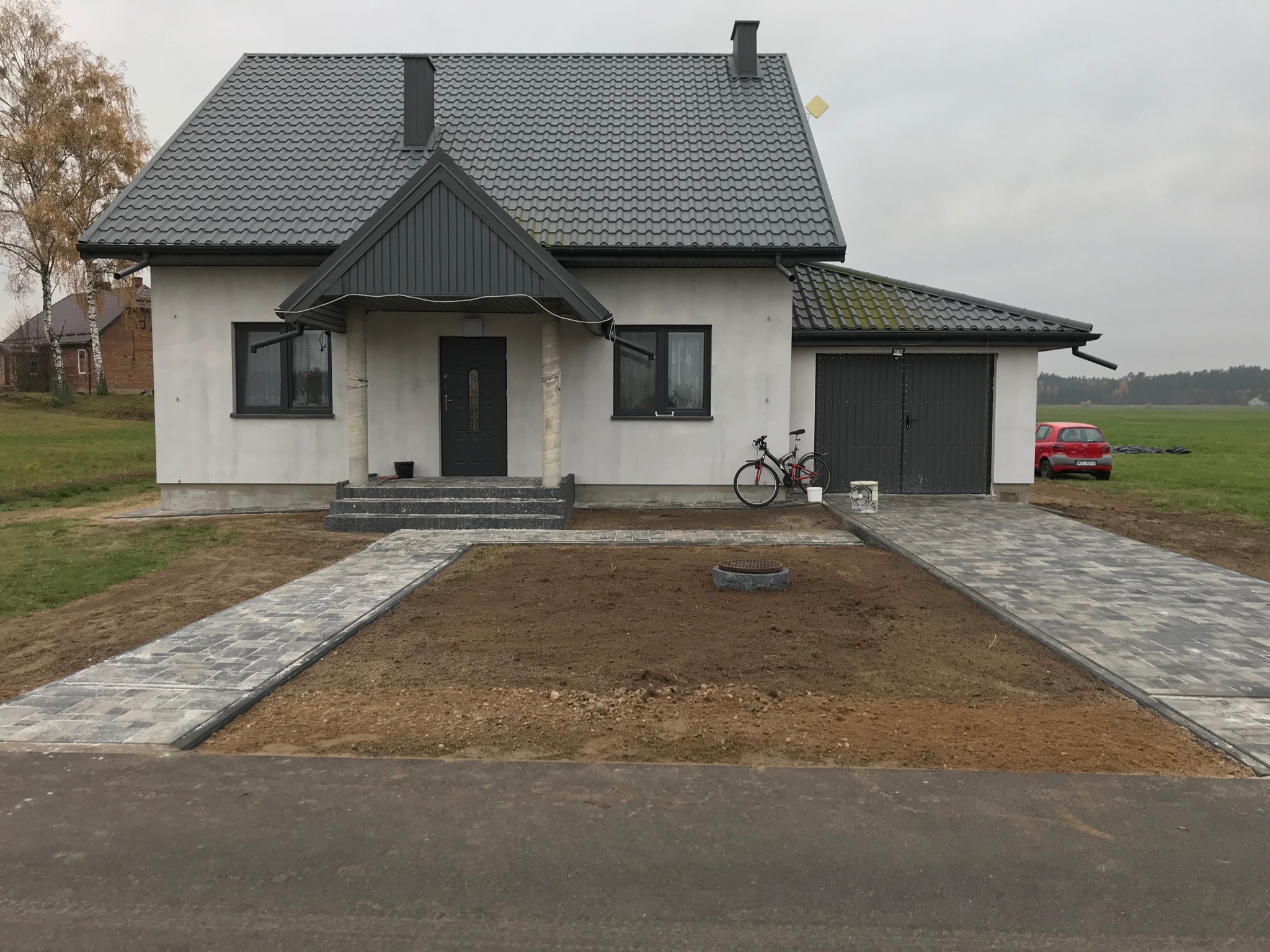 Nowy dom jednorodzinny z szarą kostką brukową na podjeździe i ścieżce do drzwi. Na podjeździe stoi rower, a w tle widać czerwony samochód i zielone pole.