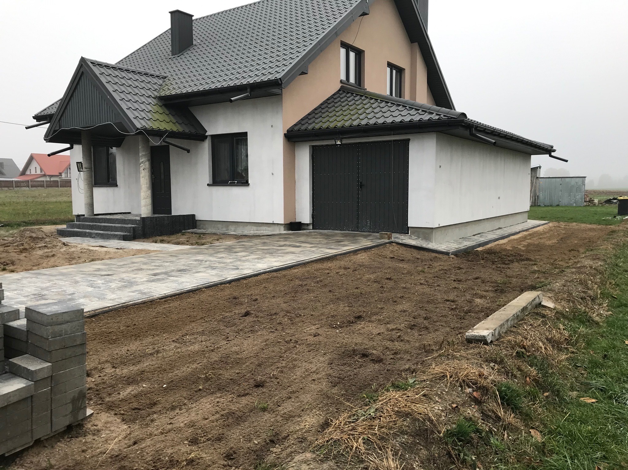 Nowy chodnik z kostki brukowej prowadzi do domu z garażem, ziemia przygotowana pod dalsze prace. Stos kostki brukowej z lewej strony.