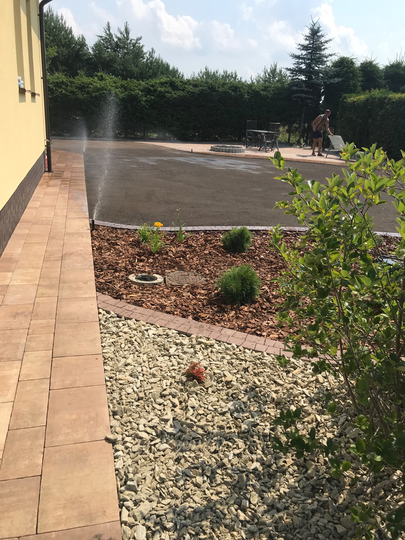 Ogród z kostką brukową, rabatami z korą i kamieniami. Automatyczne nawadnianie włączone. W tle meble ogrodowe i osoba. Słoneczny dzień.
