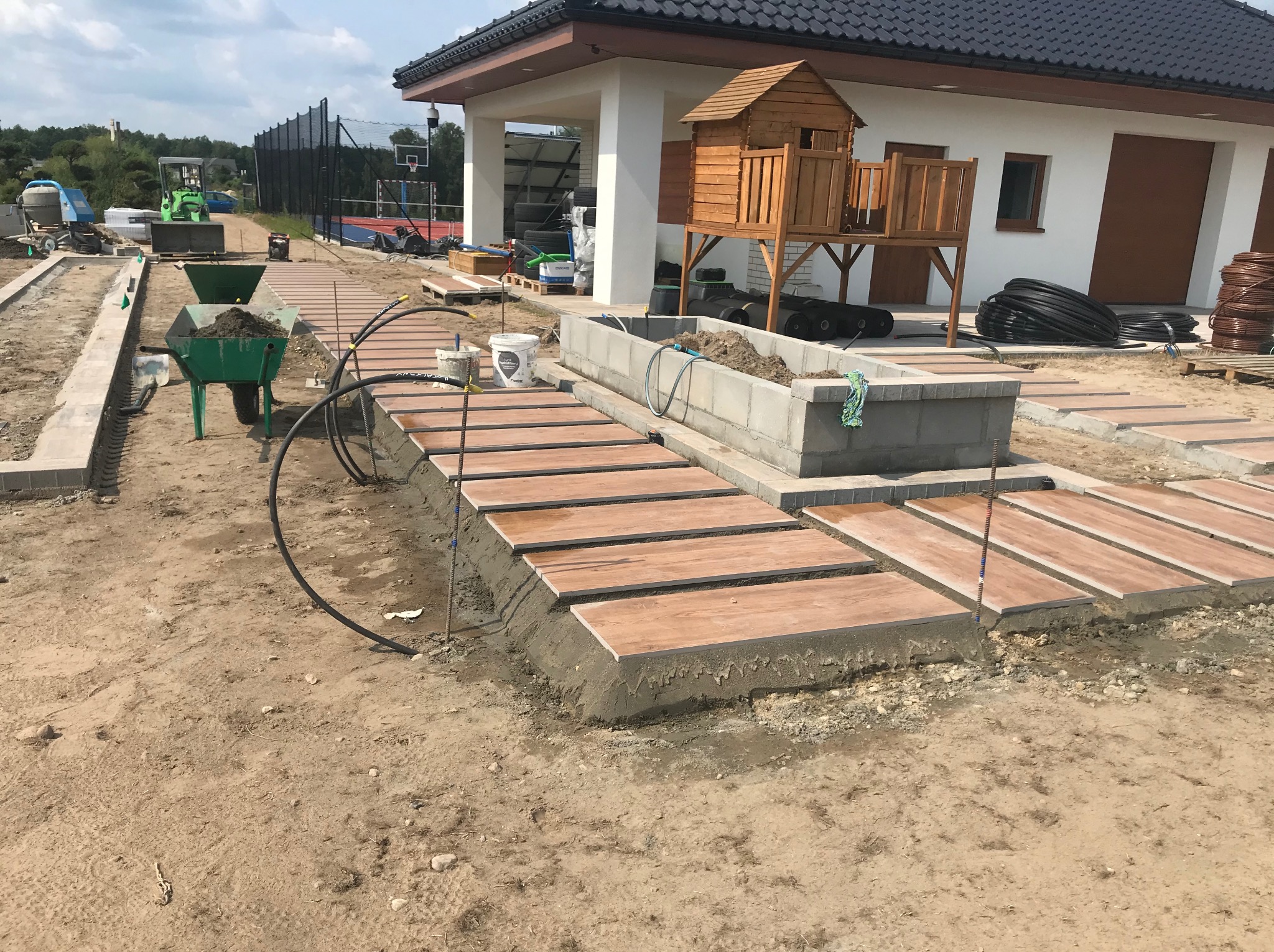 Budowa ścieżki z drewnopodobnych płyt na betonowej podstawie w ogrodzie. W tle dom z placem zabaw i sprzęt budowlany.