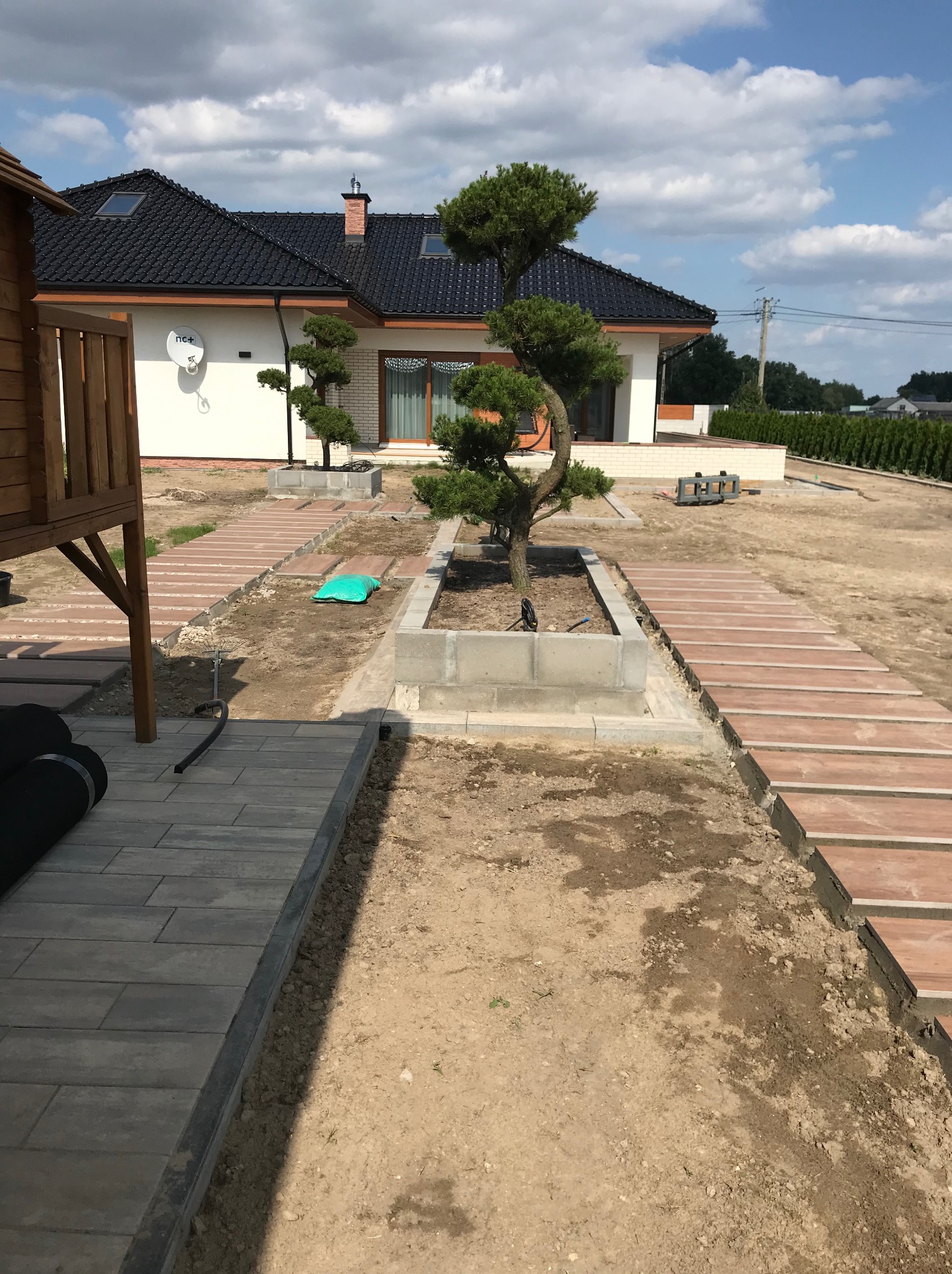 Ścieżka z kostki brukowej prowadząca do domu z ogrodem, w tle dom z czarnym dachem i białymi ścianami. Elementy małej architektury ogrodowej.