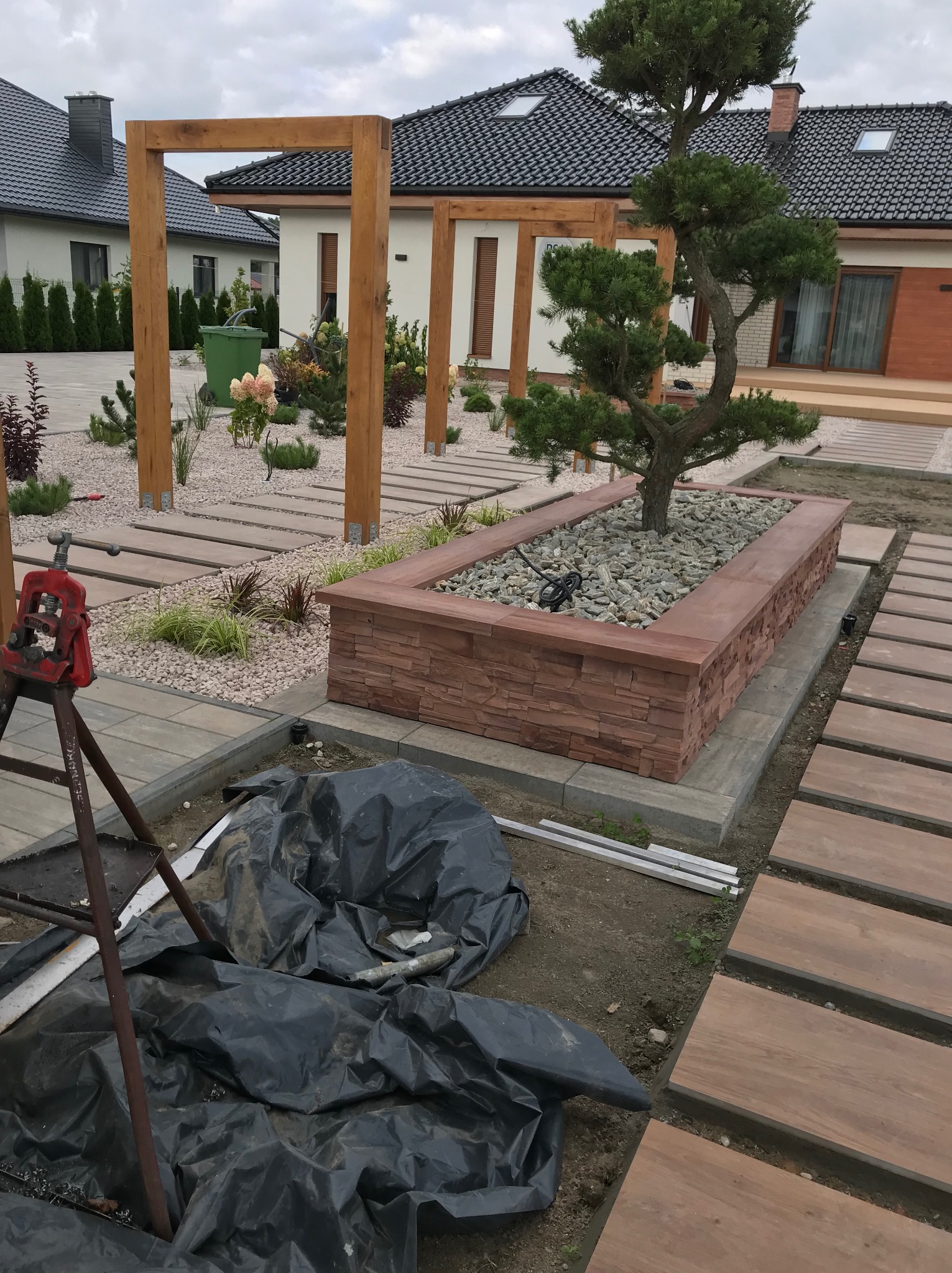 Nowoczesny ogród w trakcie realizacji: drewniane ścieżki, kamienne rabaty z drzewkiem bonsai, elementy drewnianej pergoli. W tle zabudowa jednorodzinna.