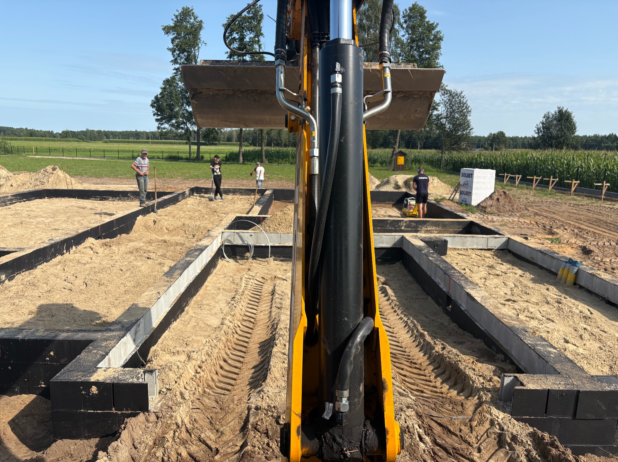 Żółta koparka w trakcie prac ziemnych przy budowie fundamentów. Widoczne szalunki i pracownicy na tle zielonego krajobrazu. Ślady opon w piasku.