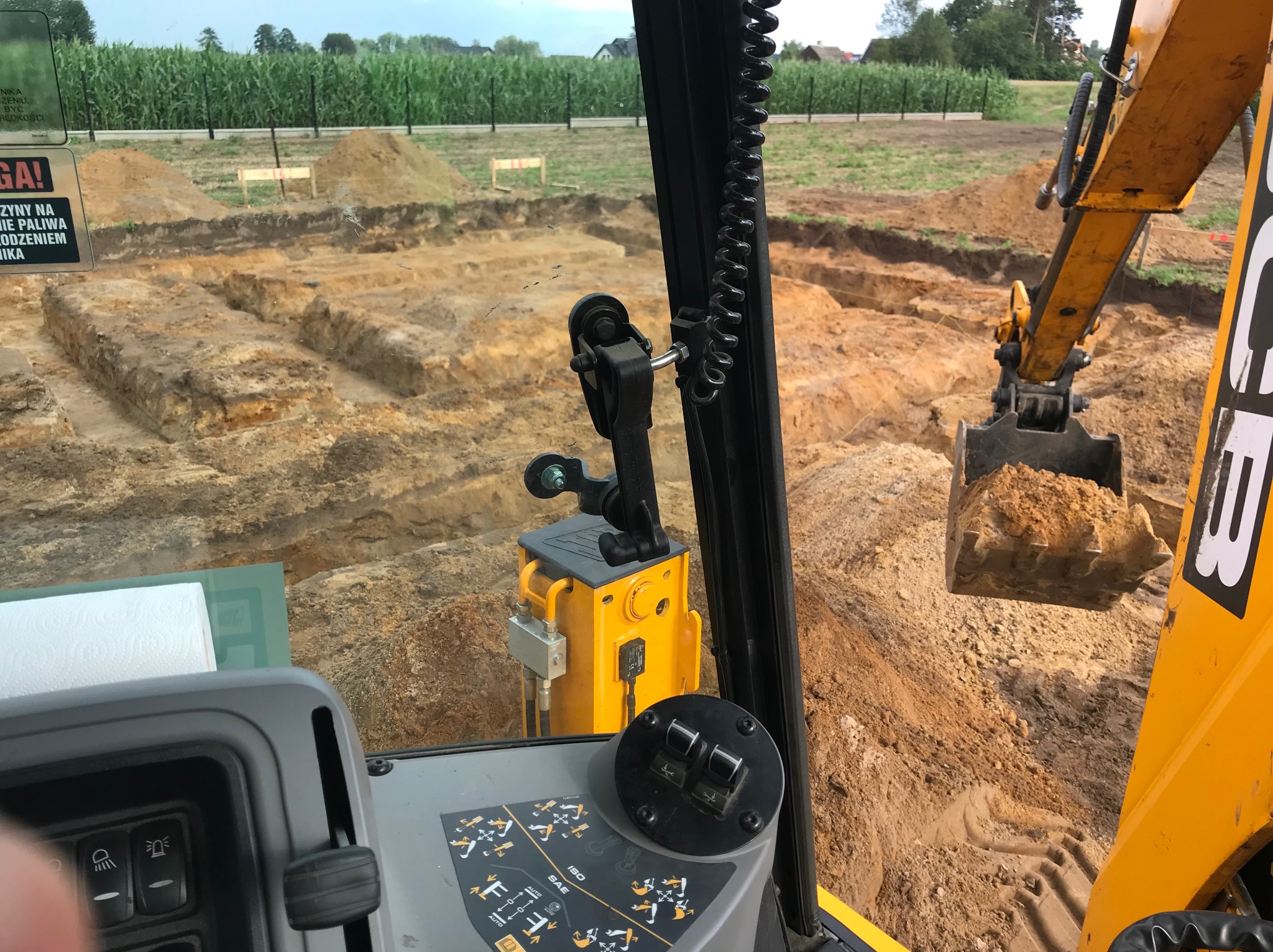 Widok z kabiny koparki na wykop pod fundamenty. Żółta maszyna w trakcie pracy na placu budowy. W tle pole kukurydzy.