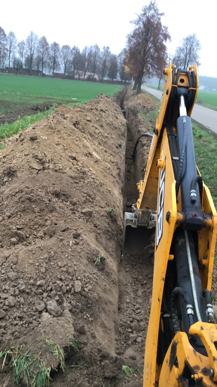 Żółta koparka JCB kopie długi, wąski wykop w ziemi, prawdopodobnie pod instalacje. W tle widoczne drzewa i łąka, co sugeruje teren podmiejski lub wiejski.