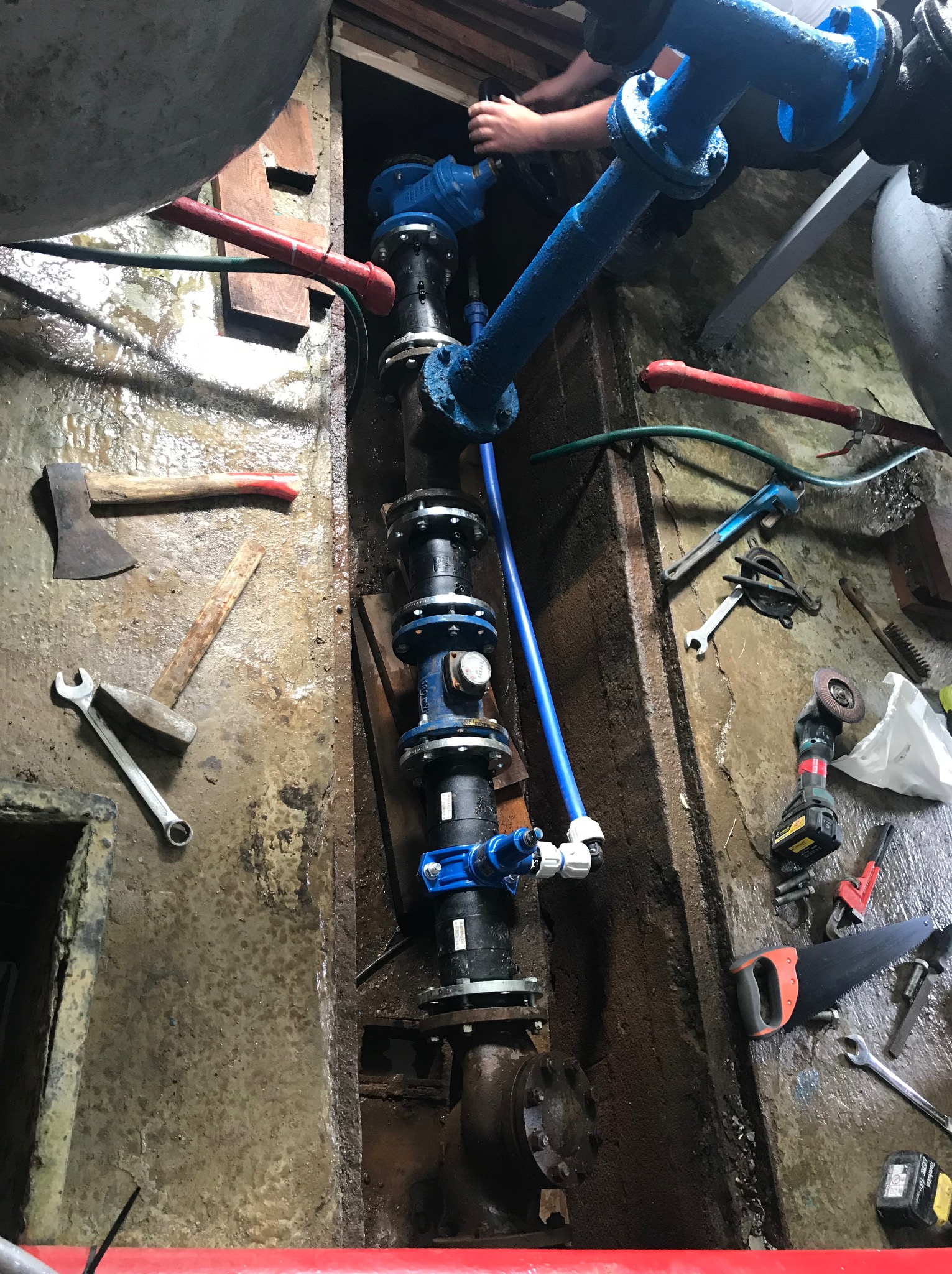 Montaż niebieskich rur w wąskim wykopie z widocznymi narzędziami: siekiera, młotek, klucze. Instalacja wodociągowa w trakcie prac, z elementami mocującymi i zaworami.