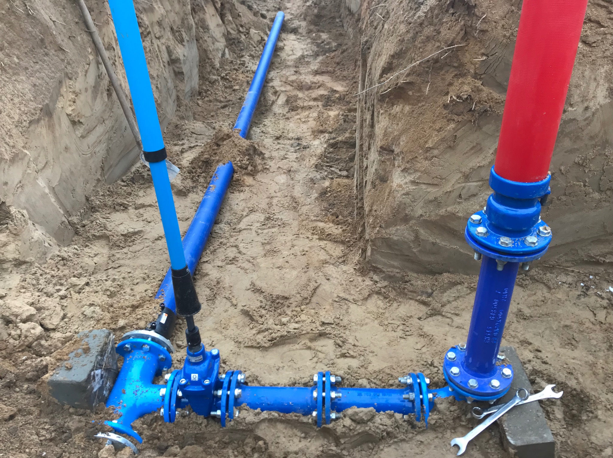 Błękitne rury wodociągowe z łącznikami w wykopie ziemnym, czerwona rura pionowa, klucze leżące obok. Instalacja wodna w trakcie montażu.