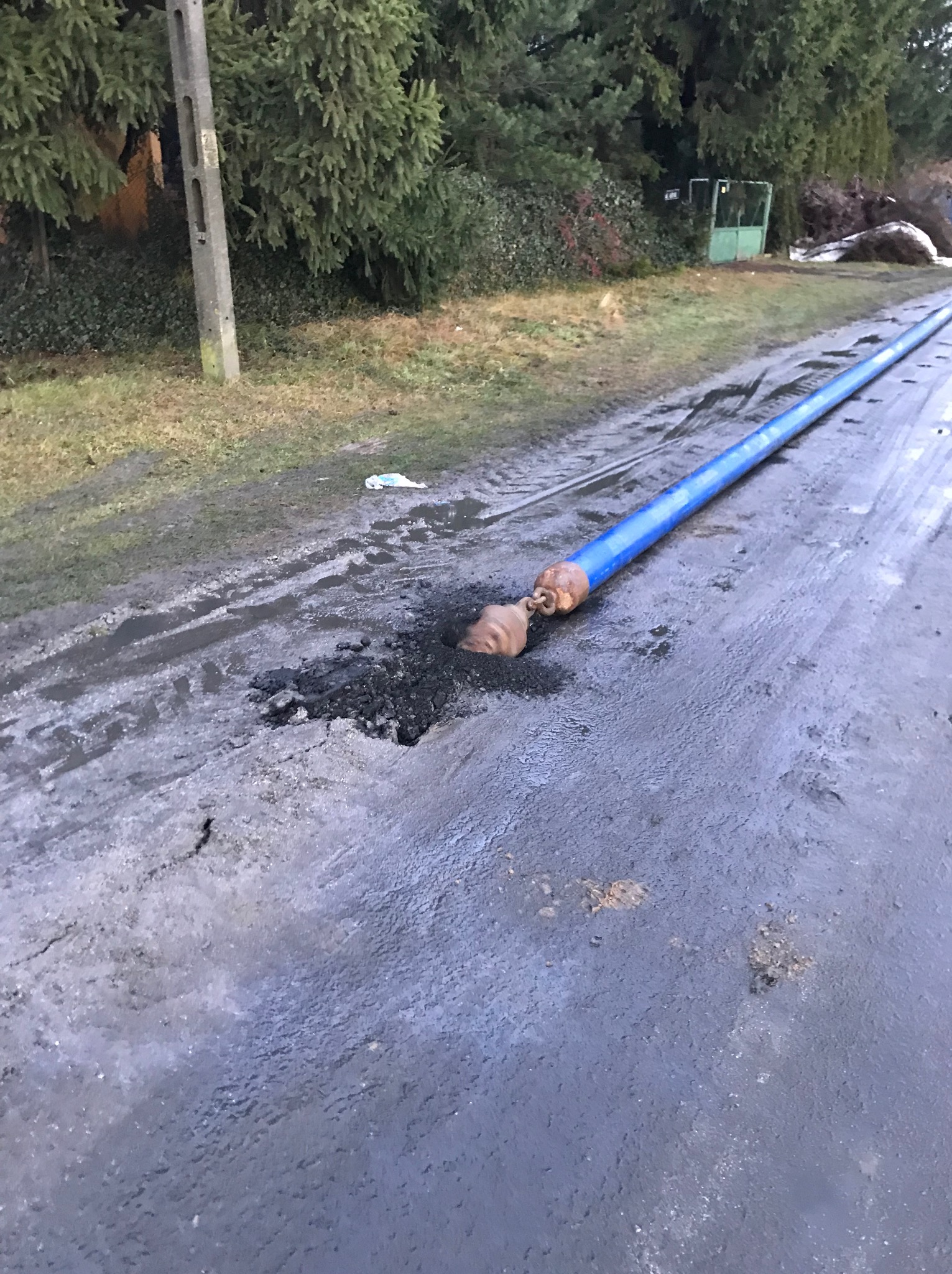 Niebieska rura z metalowym zakończeniem wystająca z wykopu na błotnistej drodze, drzewa i słup w tle. Widoczne ślady opon na drodze.