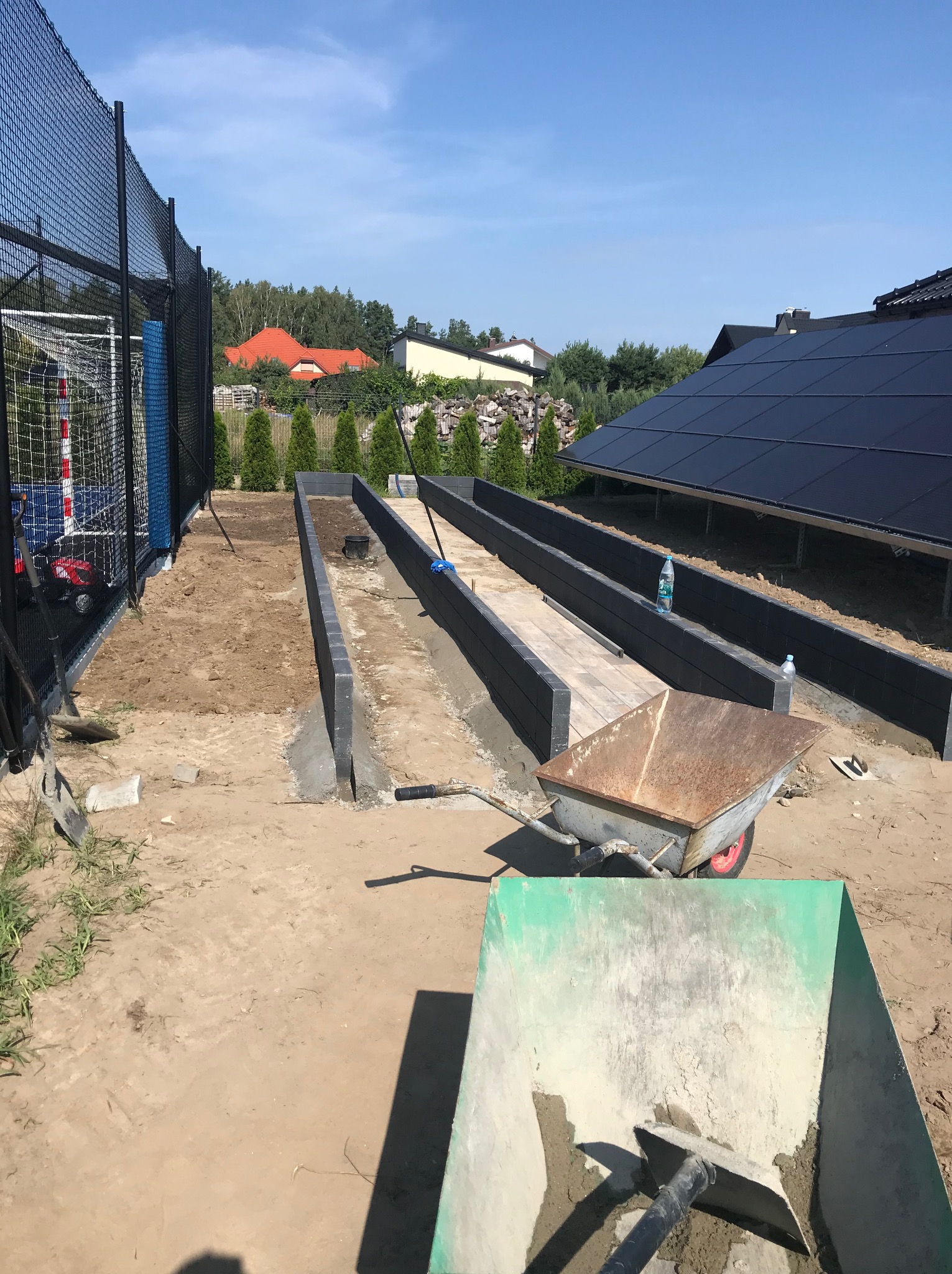 Budowa podwyższonych rabat ogrodowych z szarych elementów, widoczne narzędzia i taczki, w tle ogrodzenie boiska i panele słoneczne.