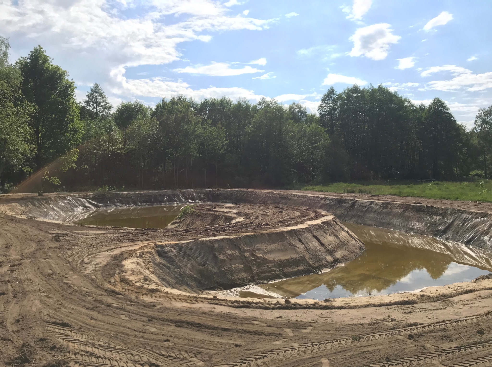 Wykonany wykop pod staw z zabezpieczoną folią skarpą i śladami opon, otoczony zielenią i błękitnym niebem z chmurami.
