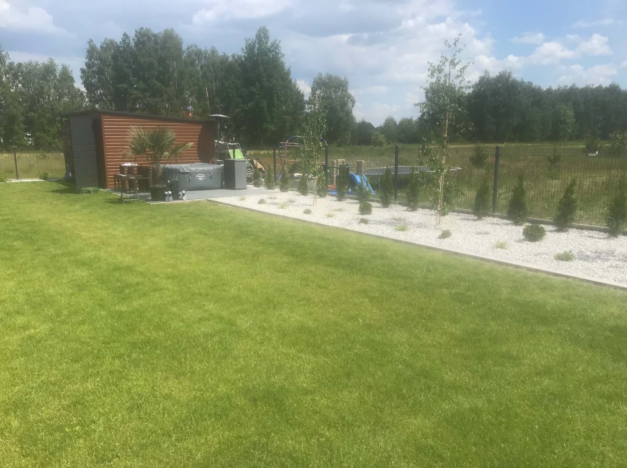 Zadbany ogród z trawnikiem, jacuzzi, domkiem narzędziowym i ogrodzeniem panelowym. Biały żwir zdobi rabaty z młodymi drzewkami. Idealne miejsce do relaksu i zabawy na świeżym powietrzu.