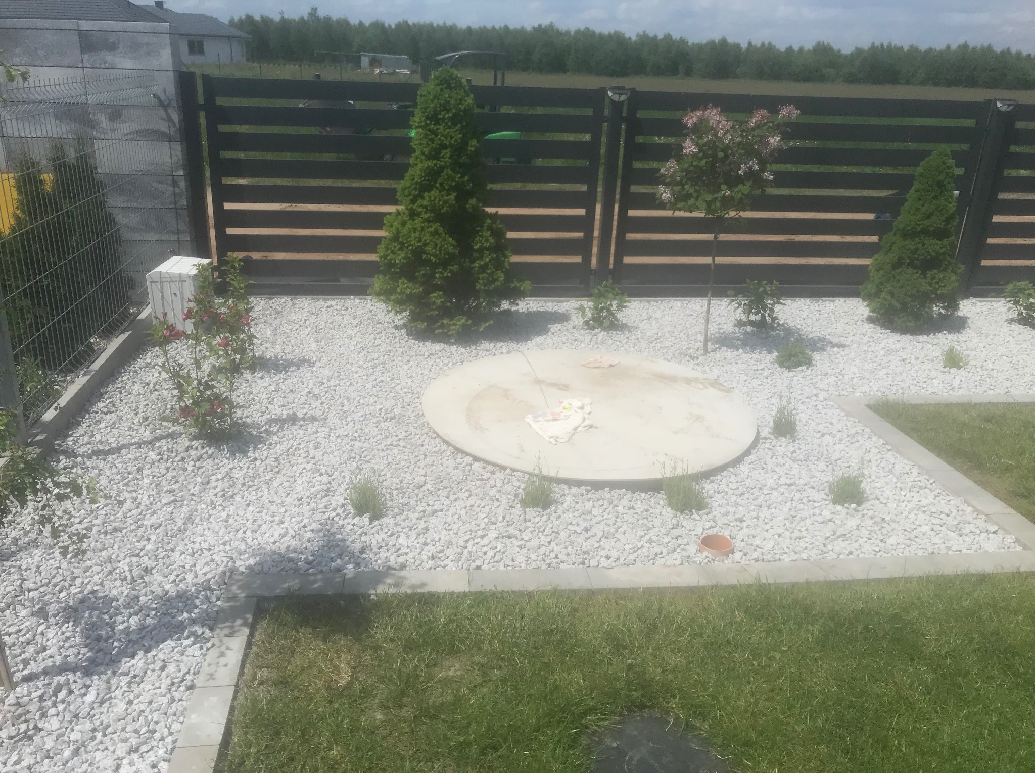 Zagospodarowany teren wokół szamba z białym grysem, obrzeżami i niską roślinnością, w tle panelowe ogrodzenie z poziomych desek i siatka.