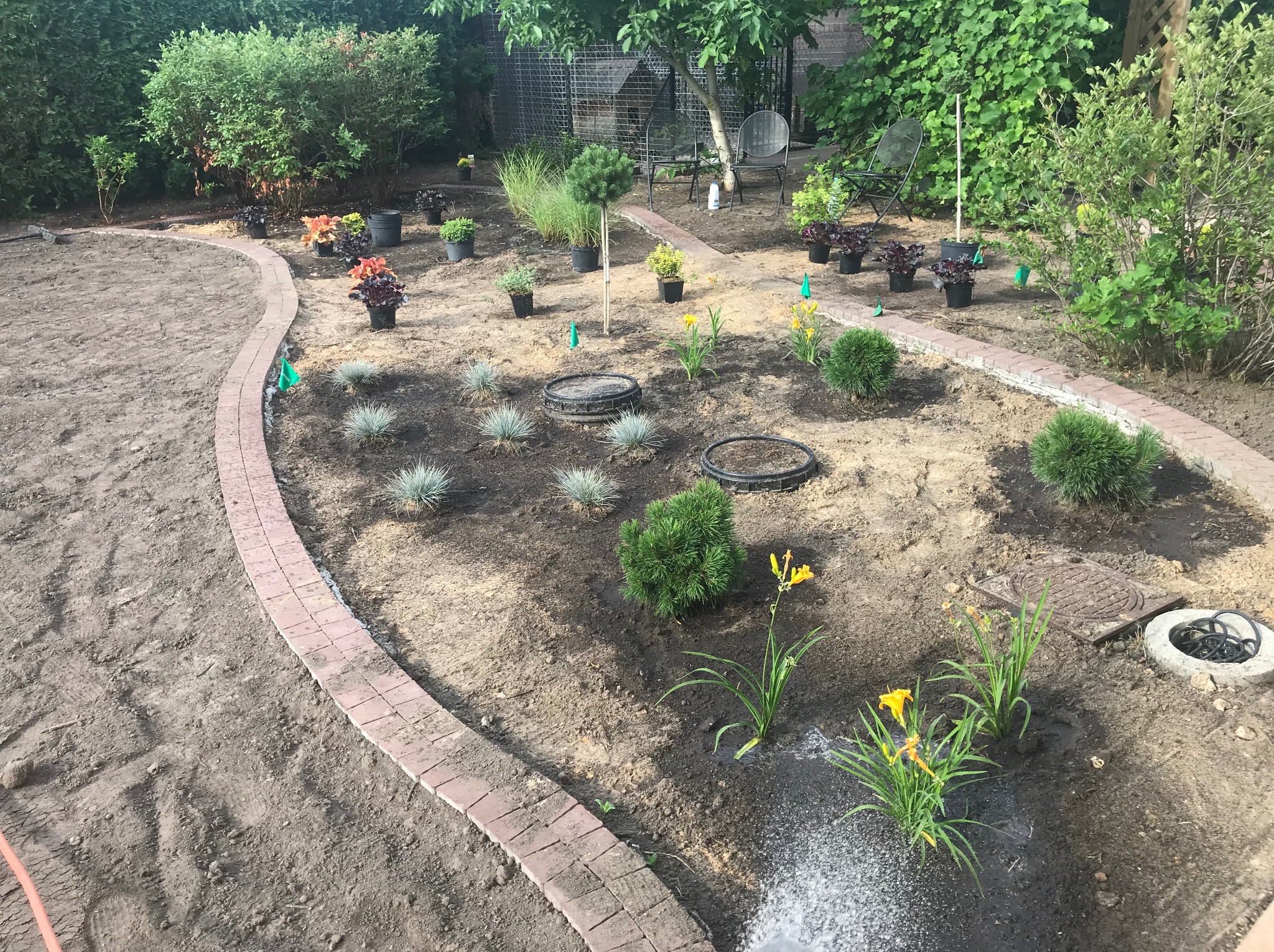 Kształtowanie ogrodu: rabata z obrzeżem z cegły, nasadzenia w donicach i ziemi, podlewanie kwiatów. W tle zielona roślinność i metalowe krzesła.