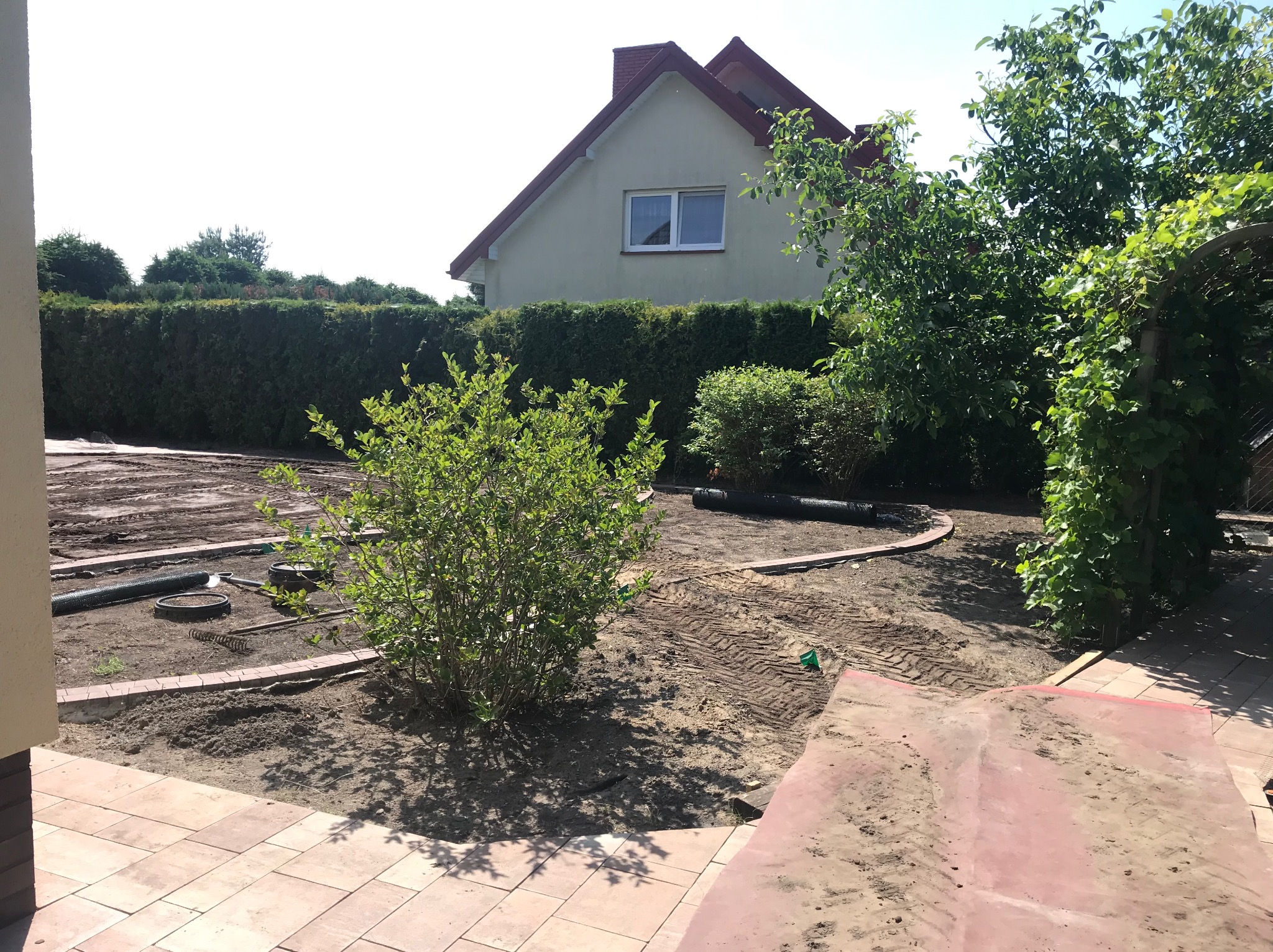 Przygotowany teren pod budowę ogrodu: wyrównana ziemia, obrzeża z kostki, narzędzia ogrodnicze. W tle dom z ogrodem i żywopłotem.
